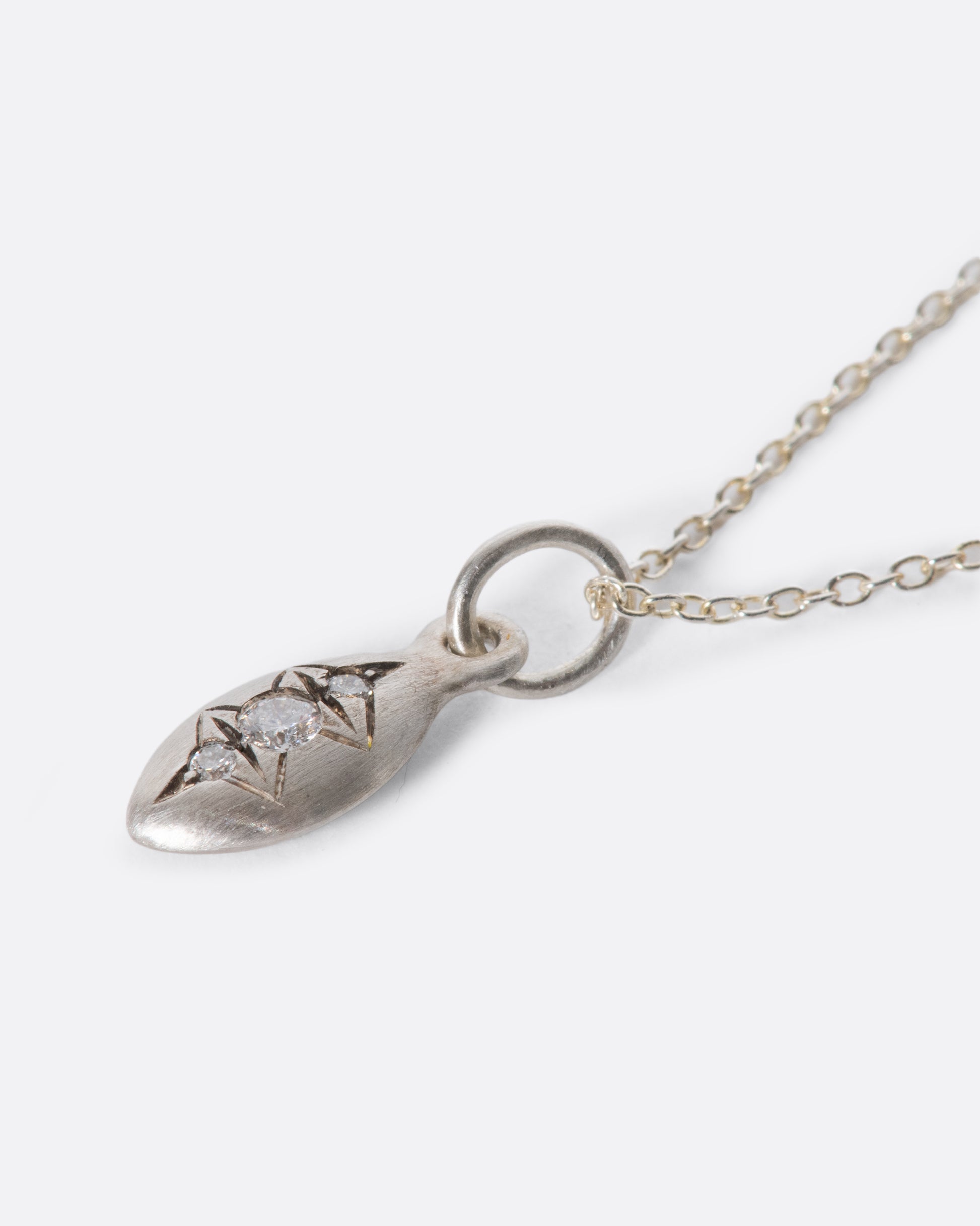 A silver cable chain necklace with a seed shaped pendant with three diamonds and oxidized etching. Shown lying flat at an angle from the side.