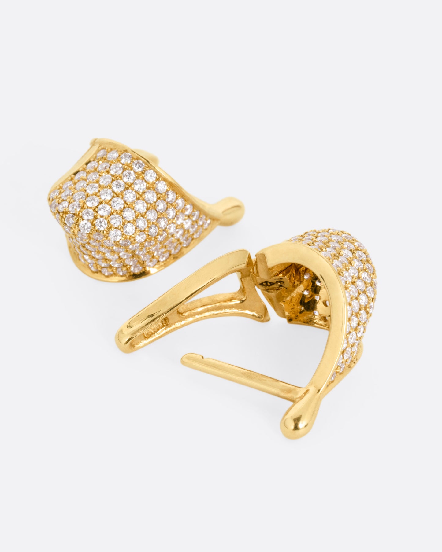 A pair of yellow gold angular hoop earrings covered in pave diamonds, shown lying on their sides with one hoop open.
