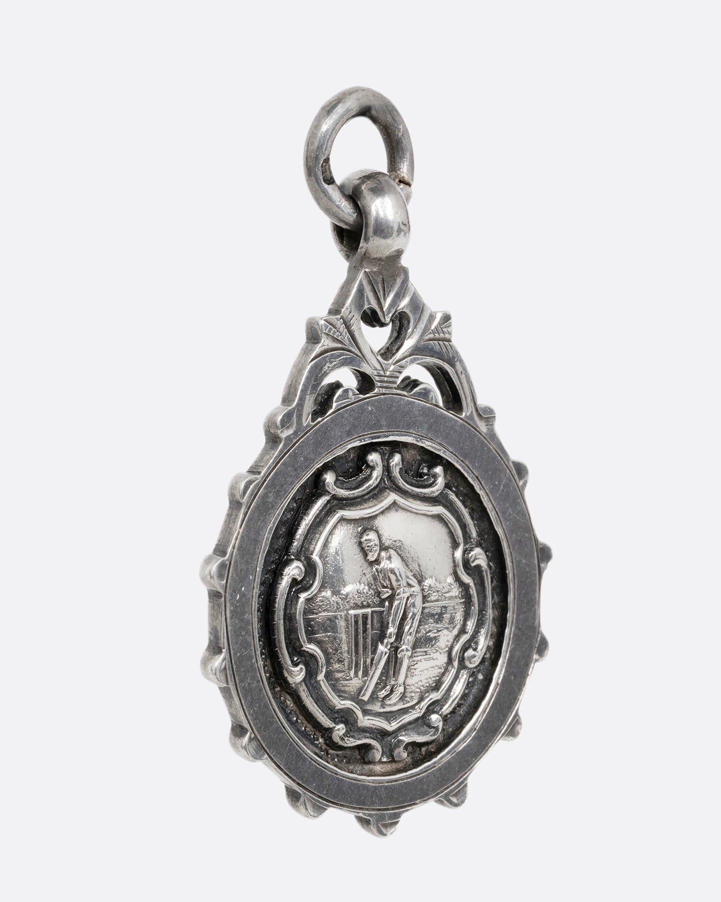 A round silver pendant with a cricket batter mid-swing surrounded by filigree detailing. Shown from the side.