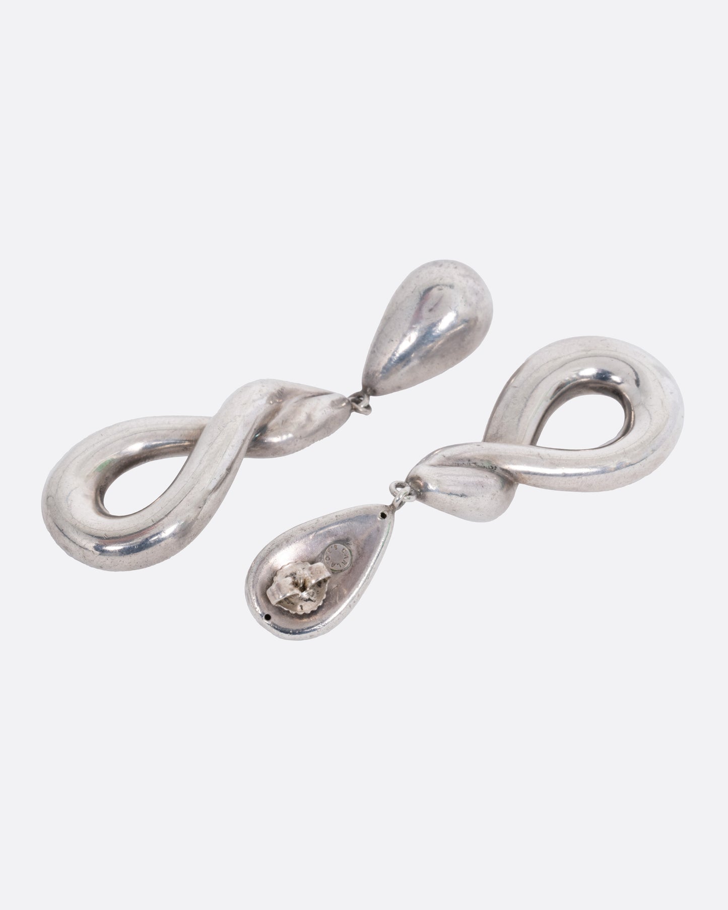A pair of silver drop earrings with a twisted silhouette. Shown lying flat- one earring from the front, the other from the back.
