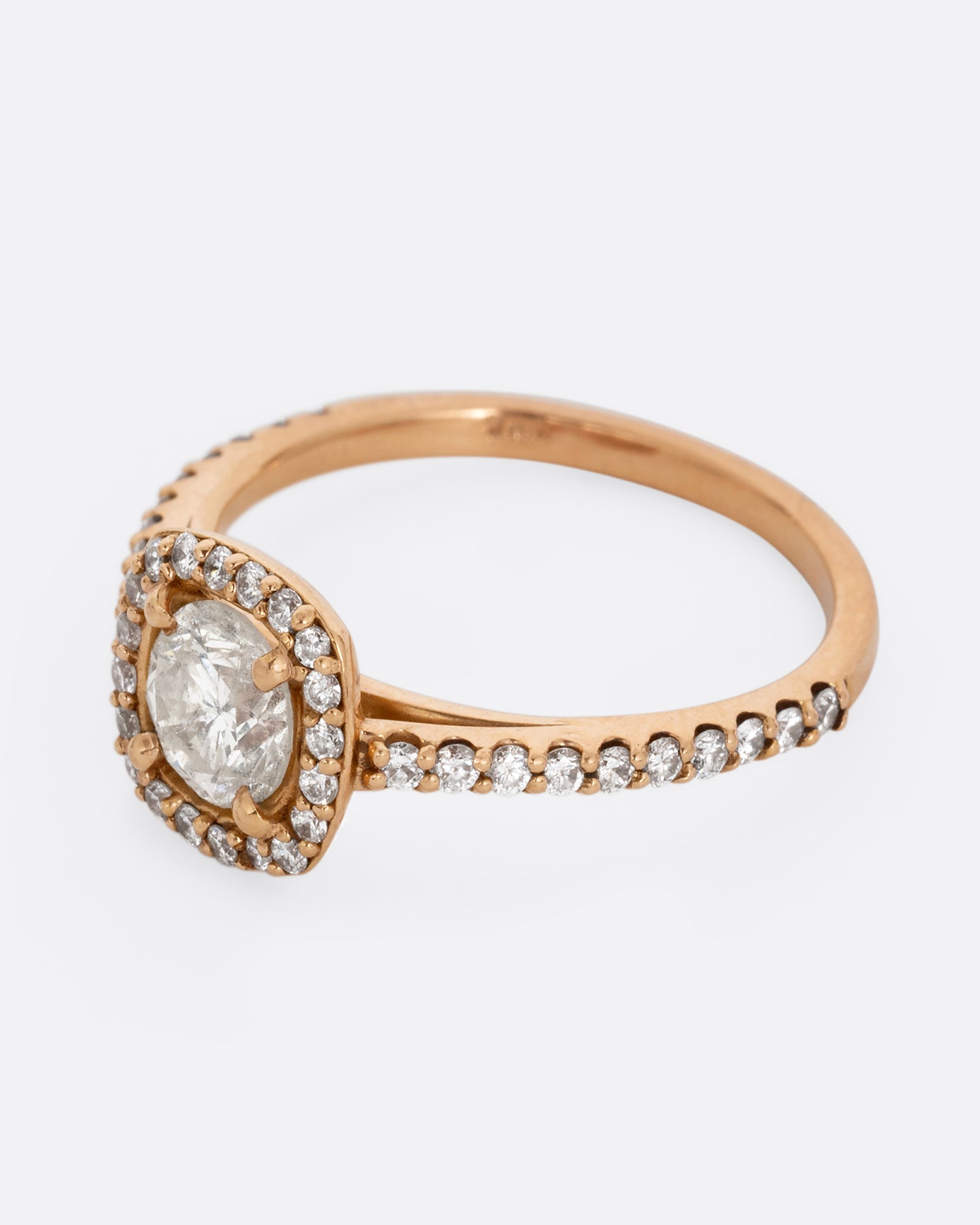 A rose gold ring with a prong set diamond surrounded by pave diamonds. The pave also wraps three quarters around the band. Shown from the side.
