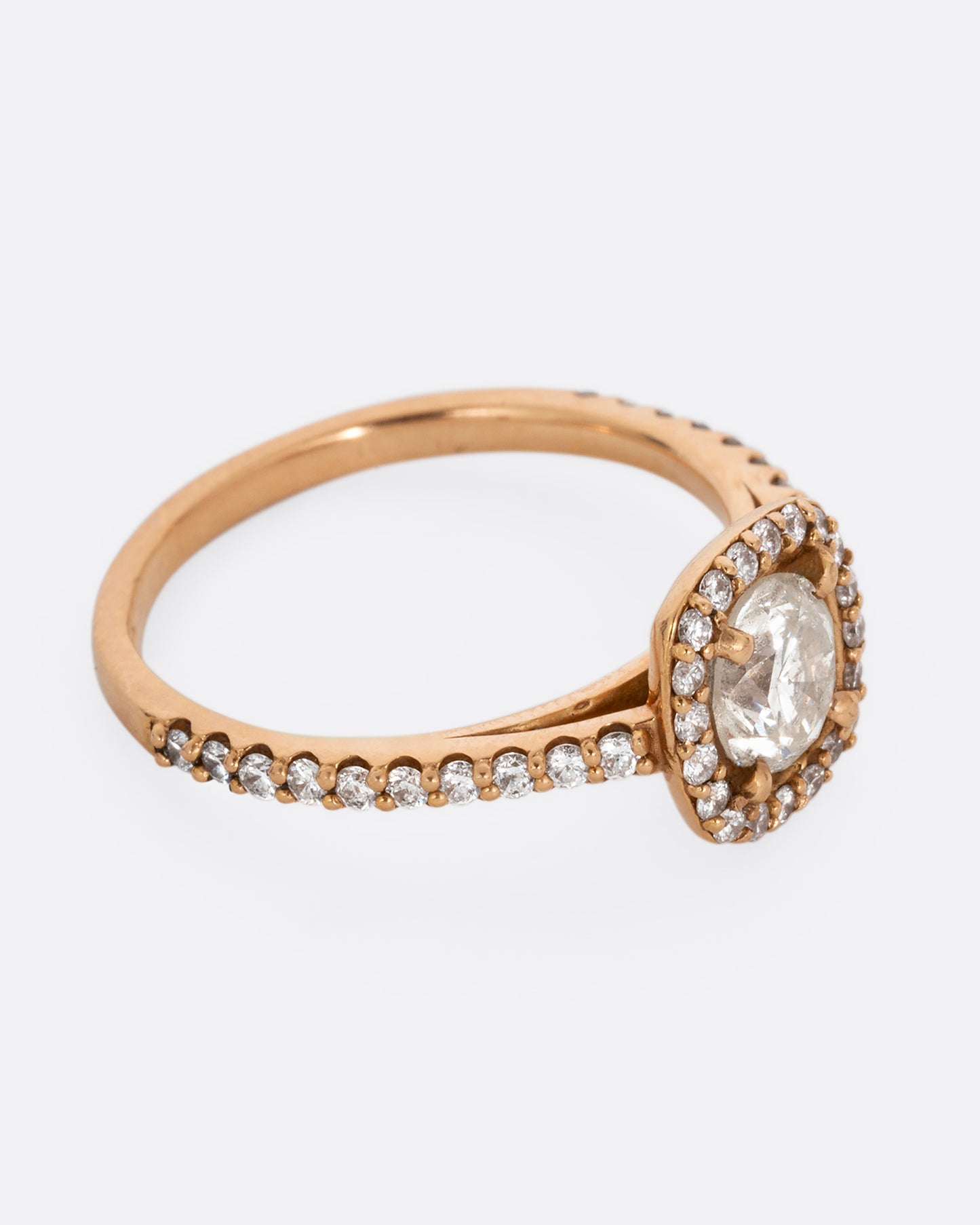 A rose gold ring with a prong set diamond surrounded by pave diamonds. The pave also wraps three quarters around the band. Shown from the side.