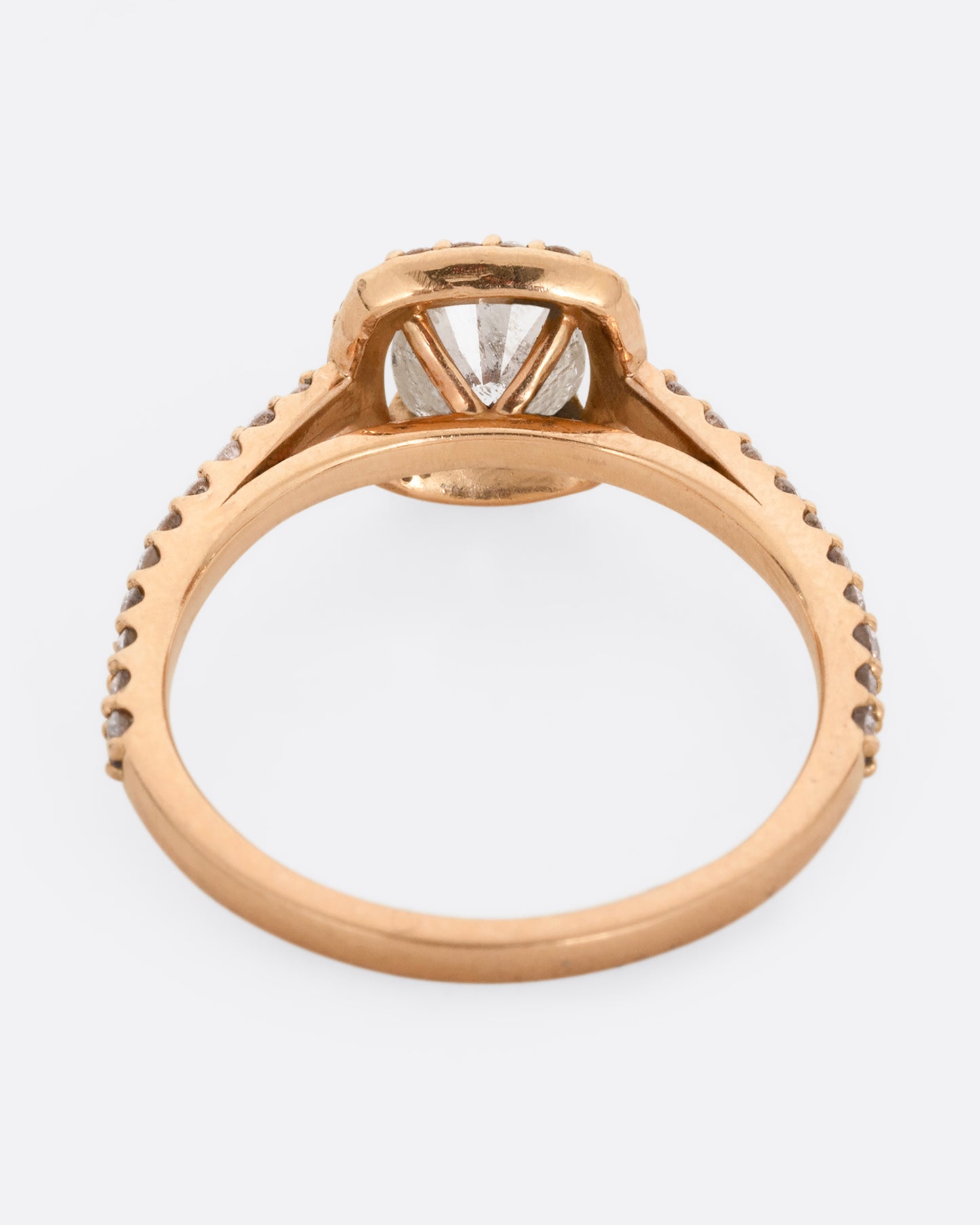 A rose gold ring with a prong set diamond surrounded by pave diamonds. The pave also wraps three quarters around the band. Shown from the back.