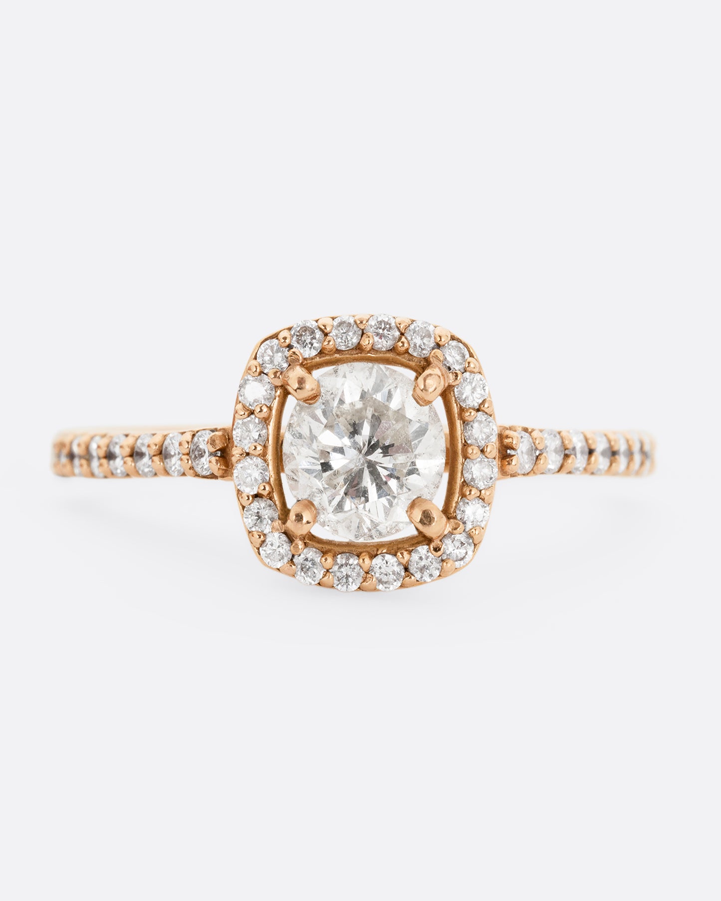 A rose gold ring with a prong set diamond surrounded by pave diamonds. The pave also wraps three quarters around the band. Shown from the front.