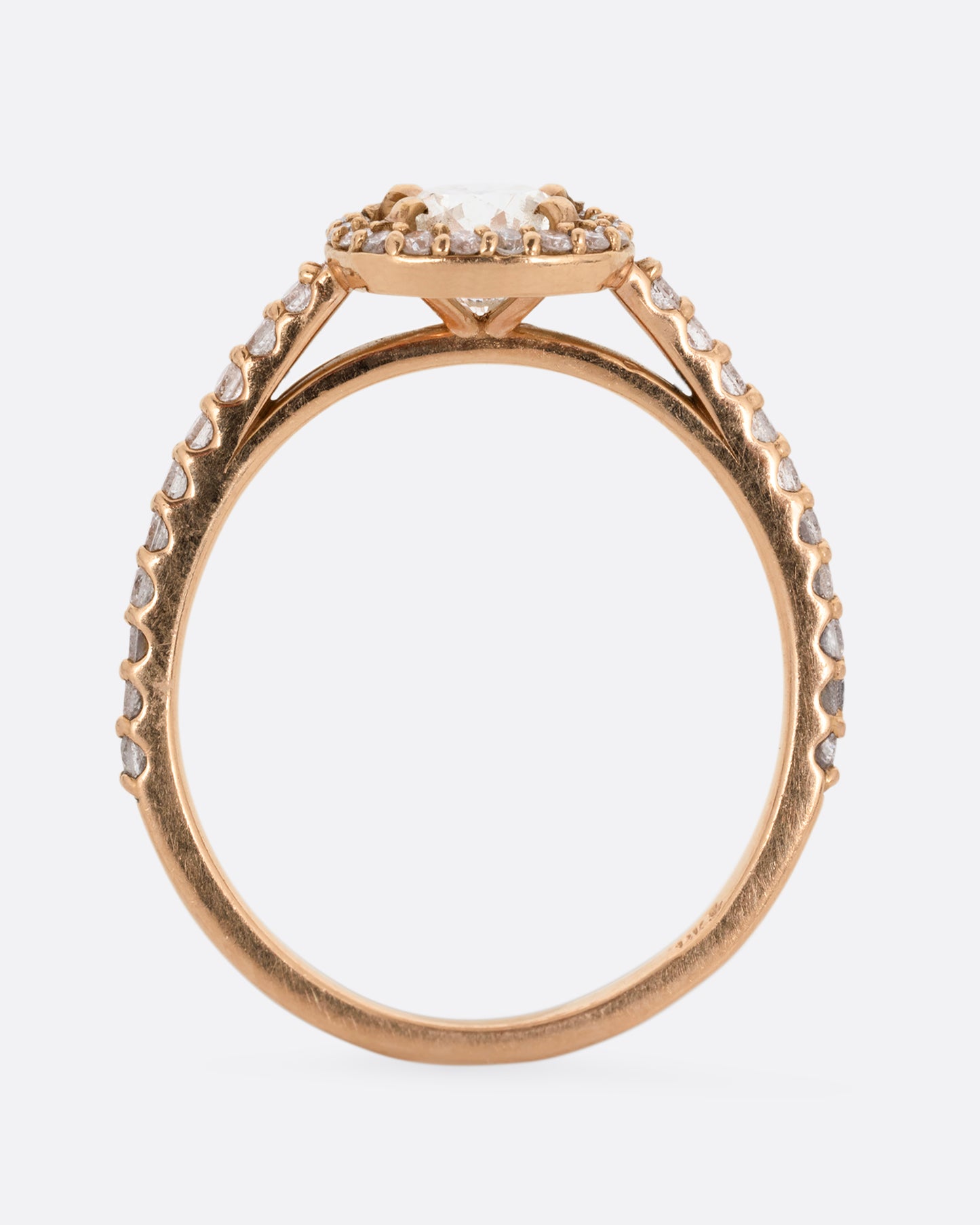 A rose gold ring with a prong set diamond surrounded by pave diamonds. The pave also wraps three quarters around the band. Shown standing up from the side.