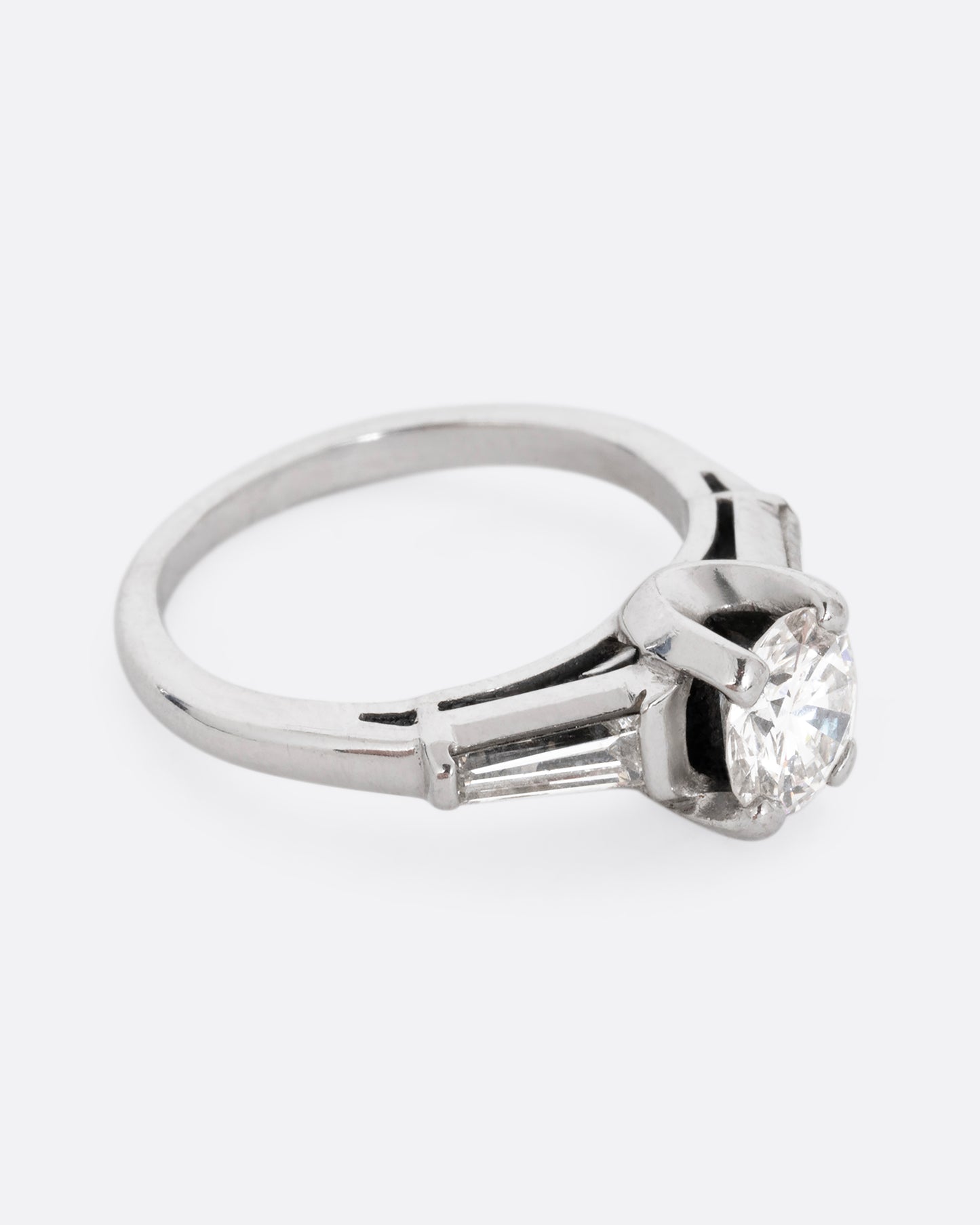 A platinum ring with a brilliant cut prong set diamond and two baguette diamonds. Shown from the side.
