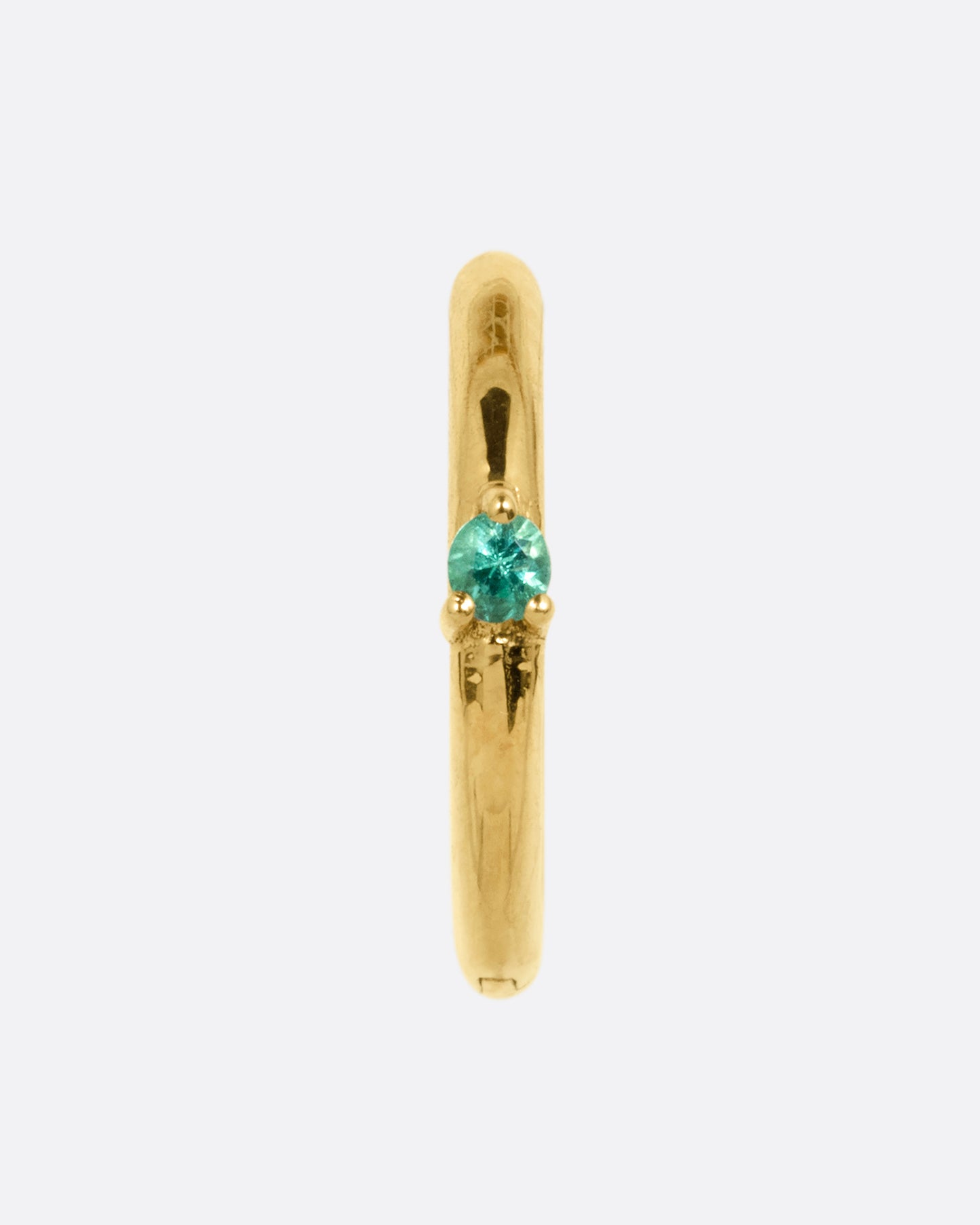 A yellow gold hinged huggie hoop with a prong set emerald at the front center. Shown from the front.