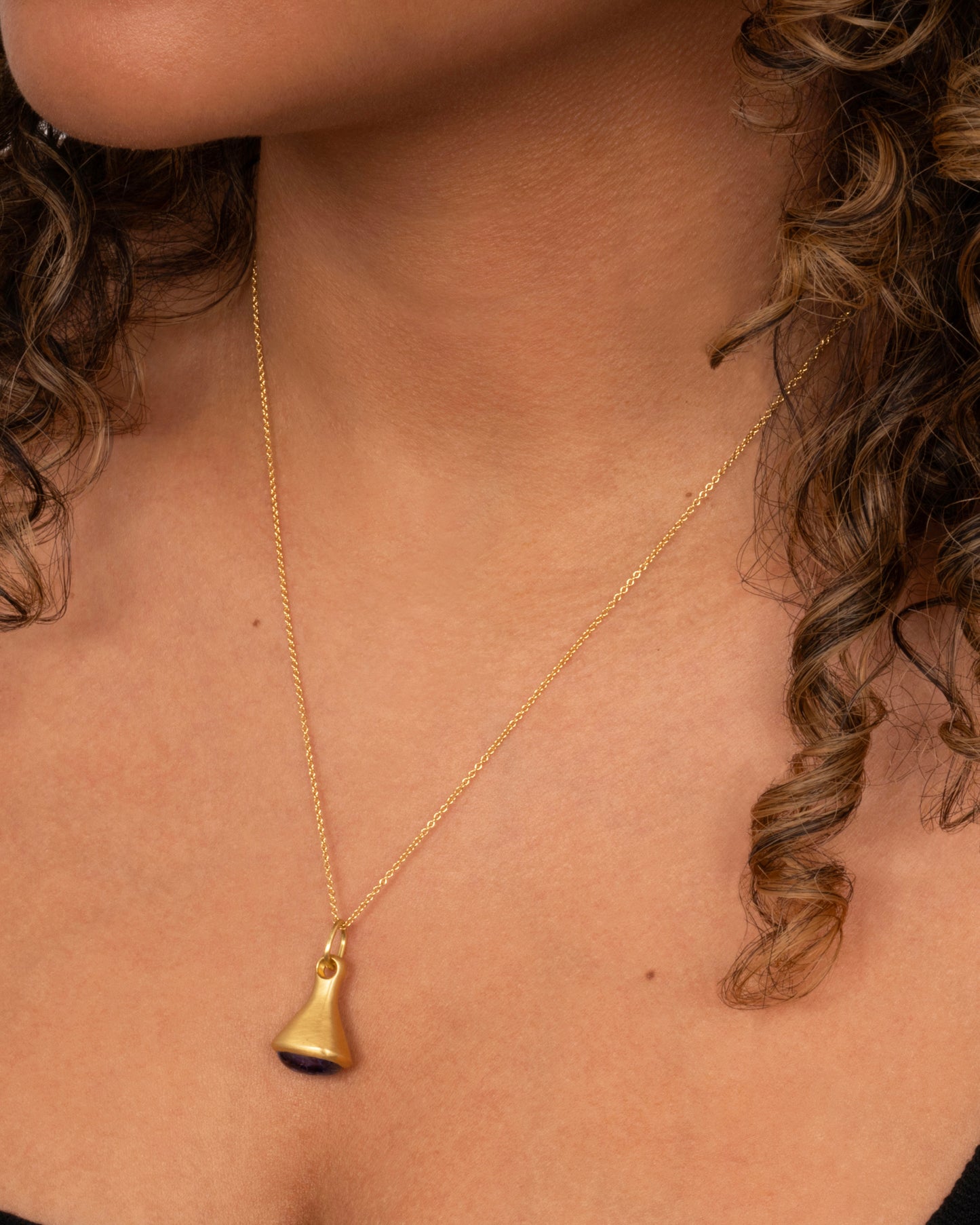 A yellow gold cable chain necklace with a bell shaped pendant with a tanzanite cabochon at the base. Shown worn around the neck.