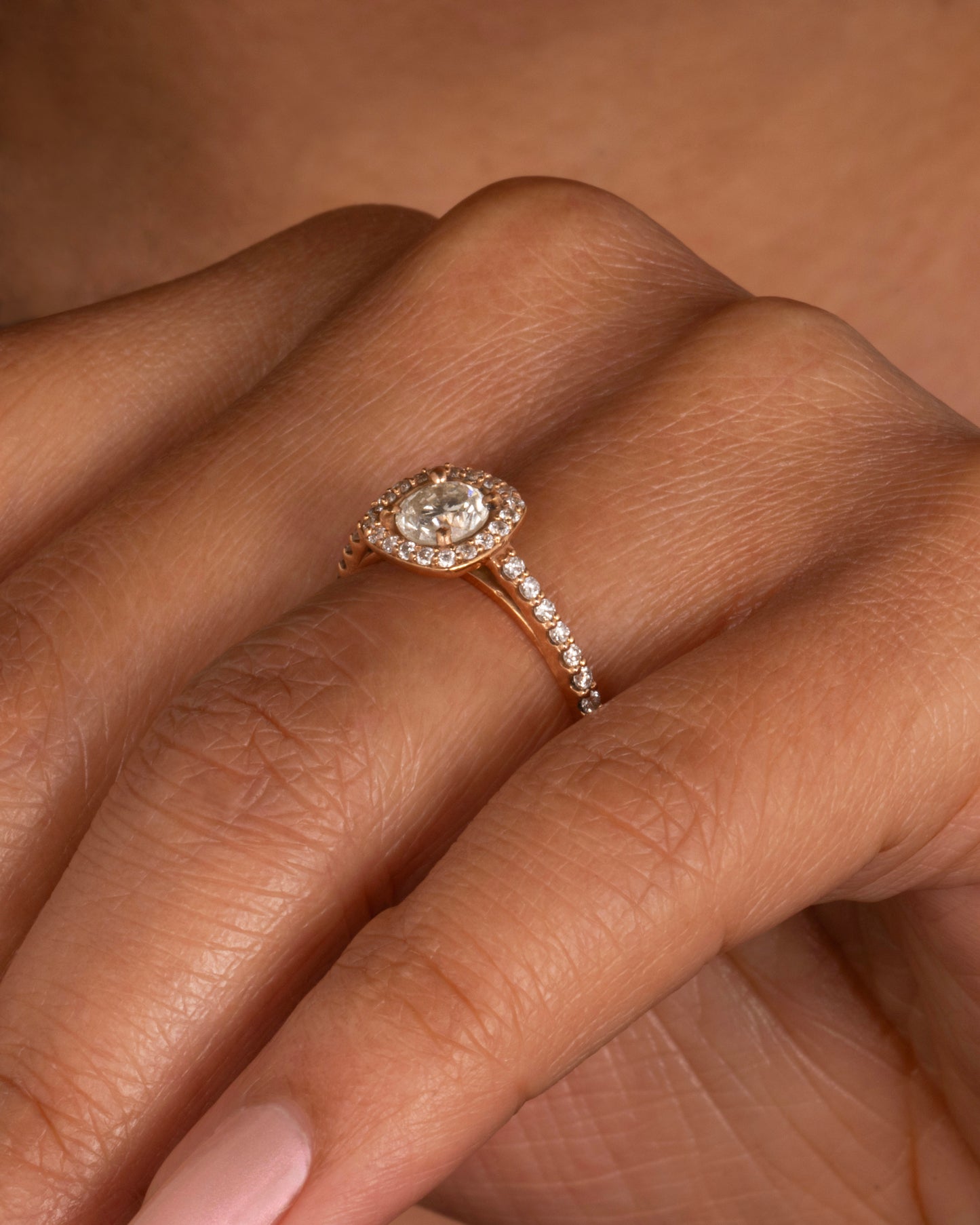 A rose gold ring with a prong set diamond surrounded by pave diamonds. The pave also wraps three quarters around the band. Shown on a hand from the side.