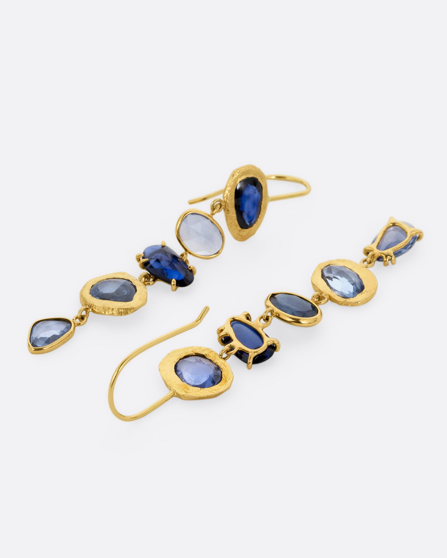 A pair of yellow gold dangle earrings with a cascade of blue rose cut sapphires with a mix of hand carved, bezel, and prong settings. One earrings is shown lying flat from the front, the other from the back.
