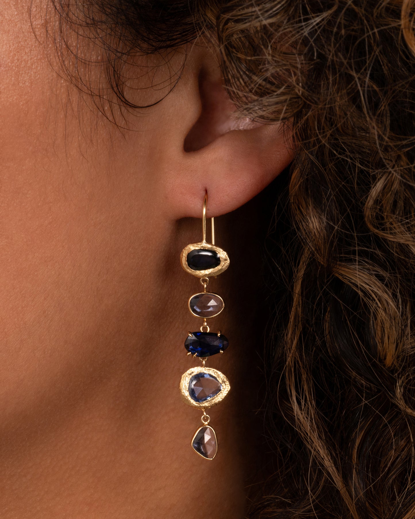 A yellow gold dangle earring with a cascade of blue rose cut sapphires with a mix of hand carved, bezel, and prong settings. Shown from the front on a person.