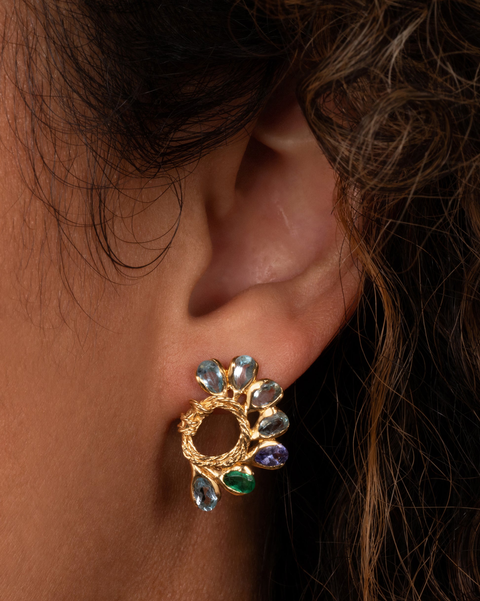 A pair of yellow gold circular twisted stud earrings feathered with oval emeralds, tanzanites, and topaz. View shown on an ear from the front.
