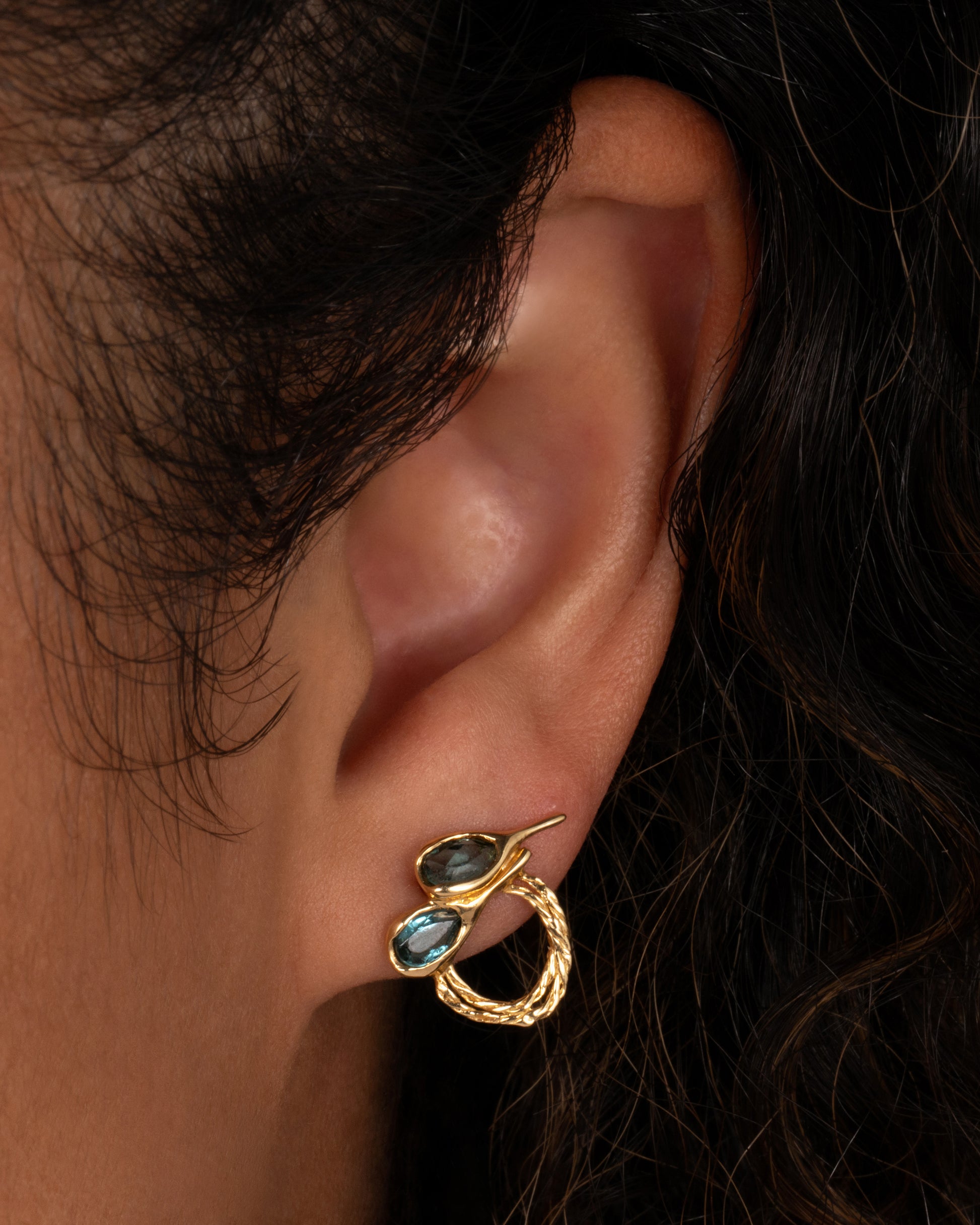 A pair of yellow gold stud earrings with two free- form blue green tourmalines and textural hoops. Shown on an ear.