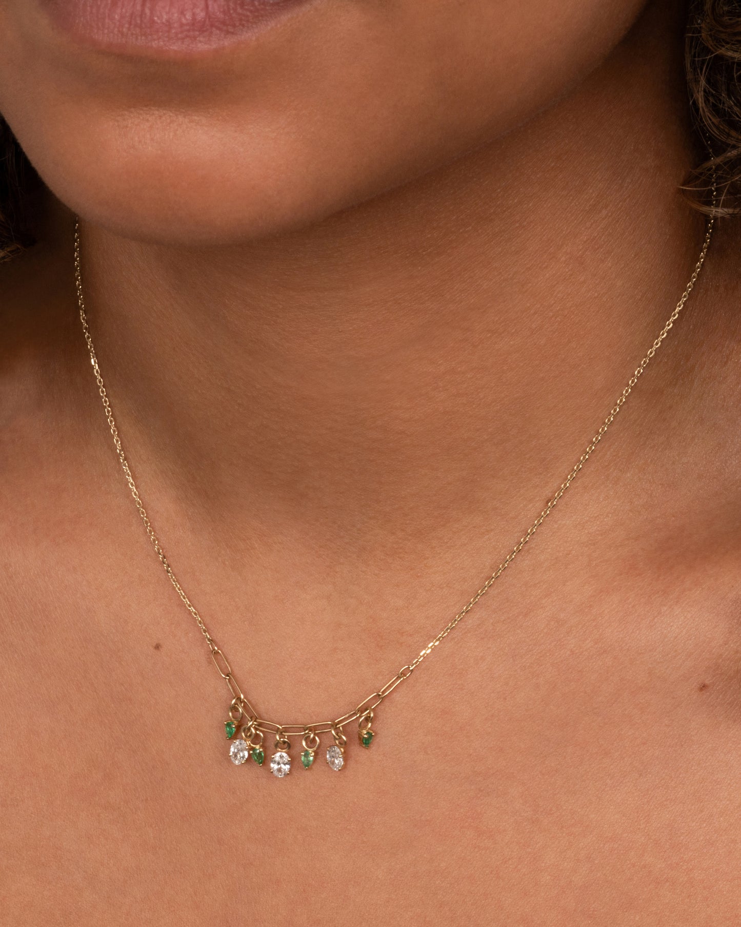 A yellow gold cable chain necklace with a paperclip chain section and diamonds and emeralds charms. Shown worn around the neck.
