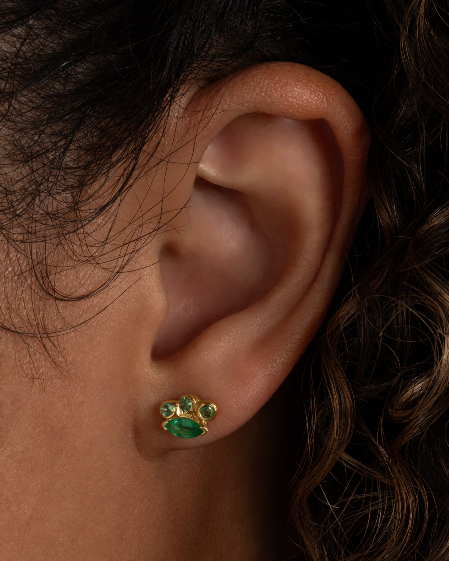 A yellow gold stud earring with round and marquise emeralds. Shown from the front on a person.