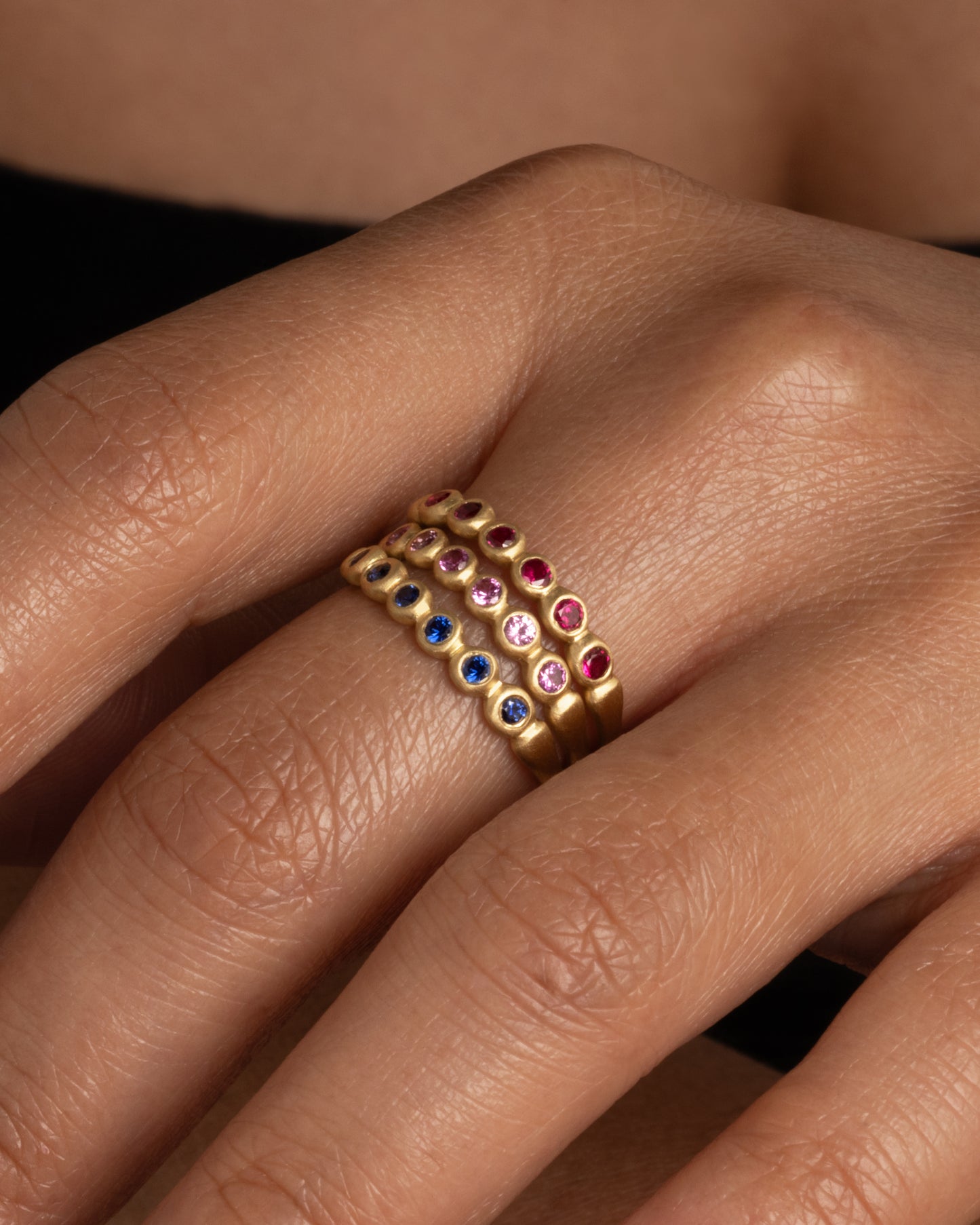 A yellow gold band with a row of pink sapphires. Shown stacked with a ruby and blue sapphire skimmer band worn on a finger.