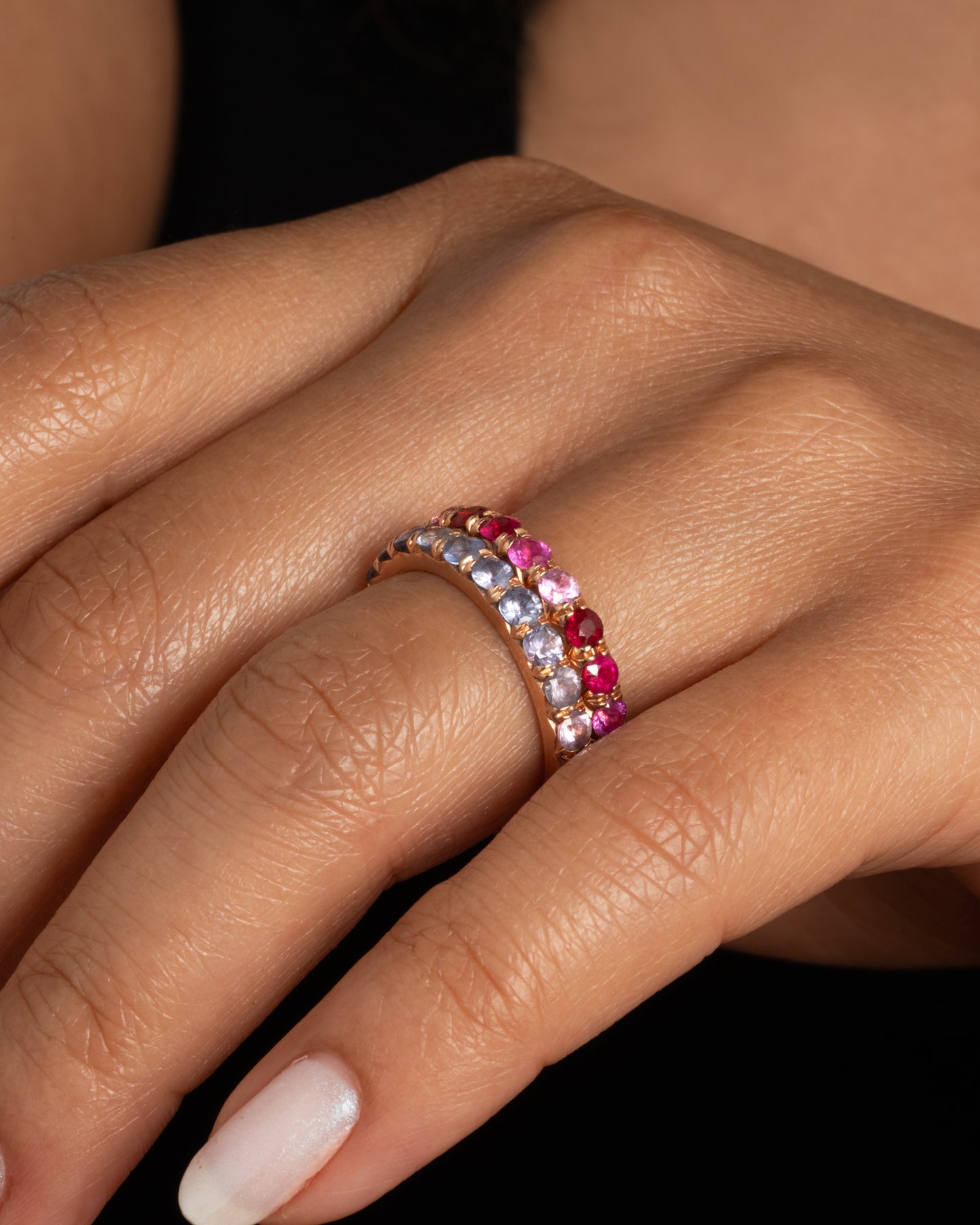 A rose gold eternity band with pink and blue cut sapphires. Shown paired with another Tamsin band featuring reds and pinks worn on a finger.