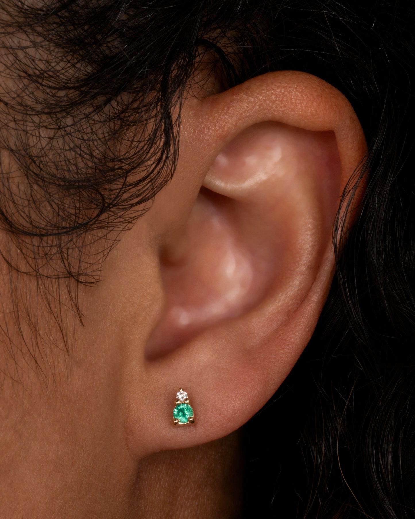 A yellow gold stud earring with a diamond on top and an emerald on the bottom. Shown from the front on a person.