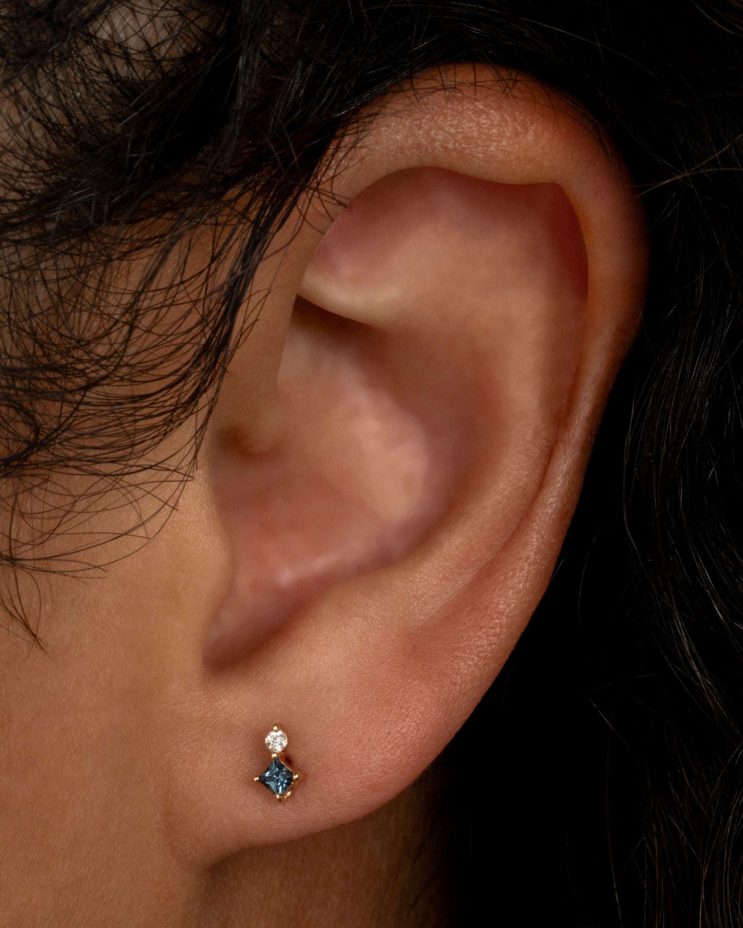 A yellow gold stud earring with a prong set round diamond sitting atop a princess cut teal blue sapphire. Shown worn in an ear.