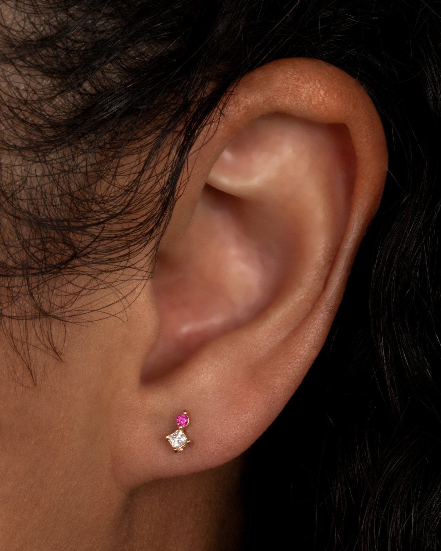 A yellow gold stud earring with a princess cut diamond and a round pink sapphire. Shown from the front on a person.