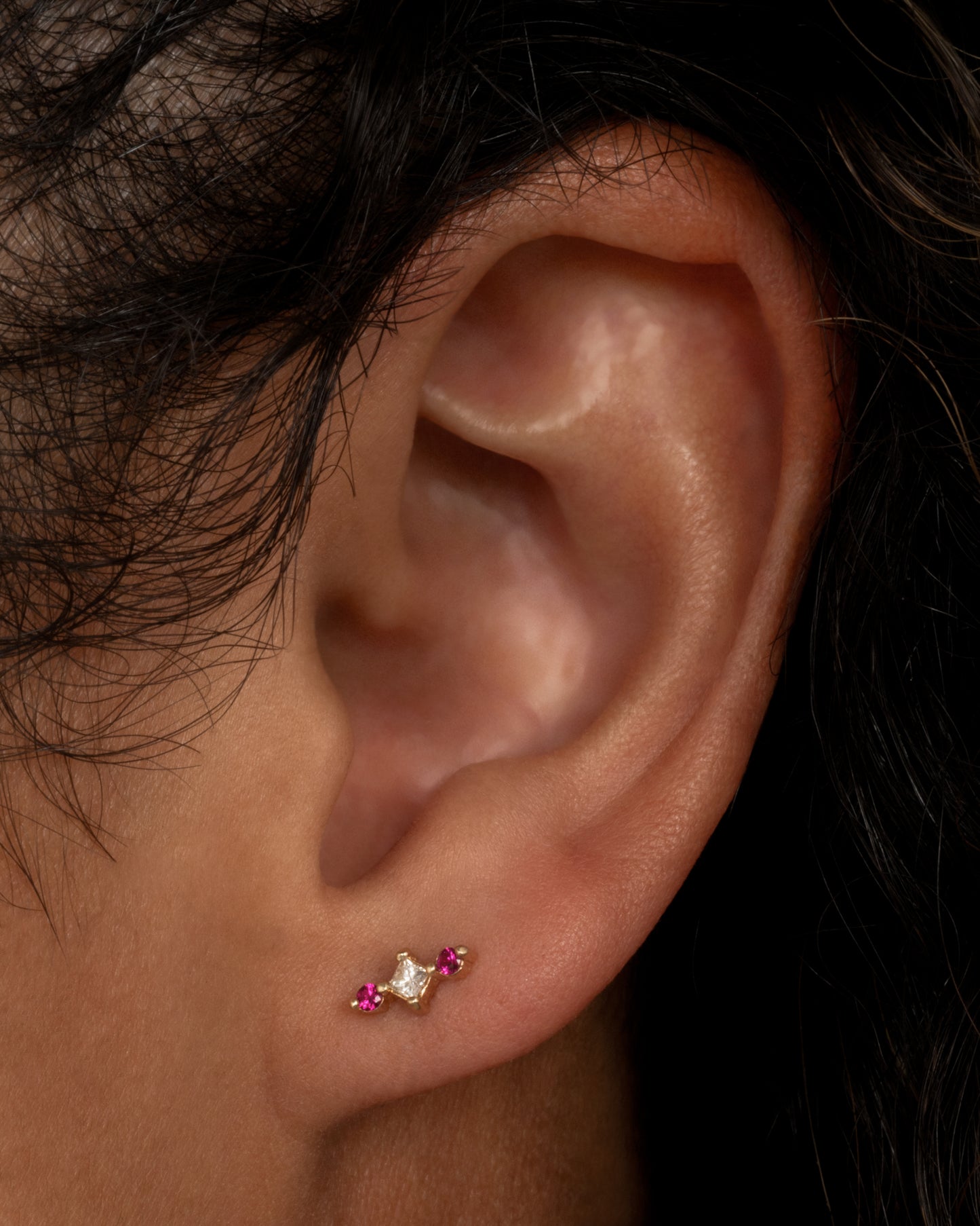 A yellow gold stud earring with a princess cut diamond at the center and two round pink sapphires on either side. Shown from the front worn in an ear.