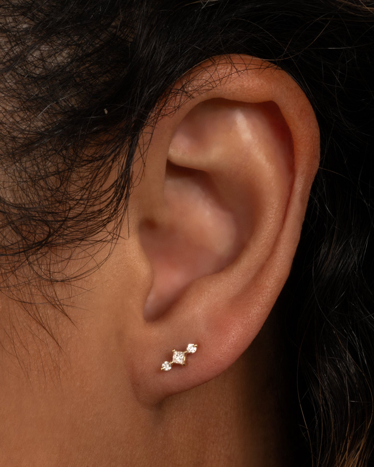 A white gold stud earring with a princess cut diamond at the center and two round diamonds on either side. View from the front on a person.
