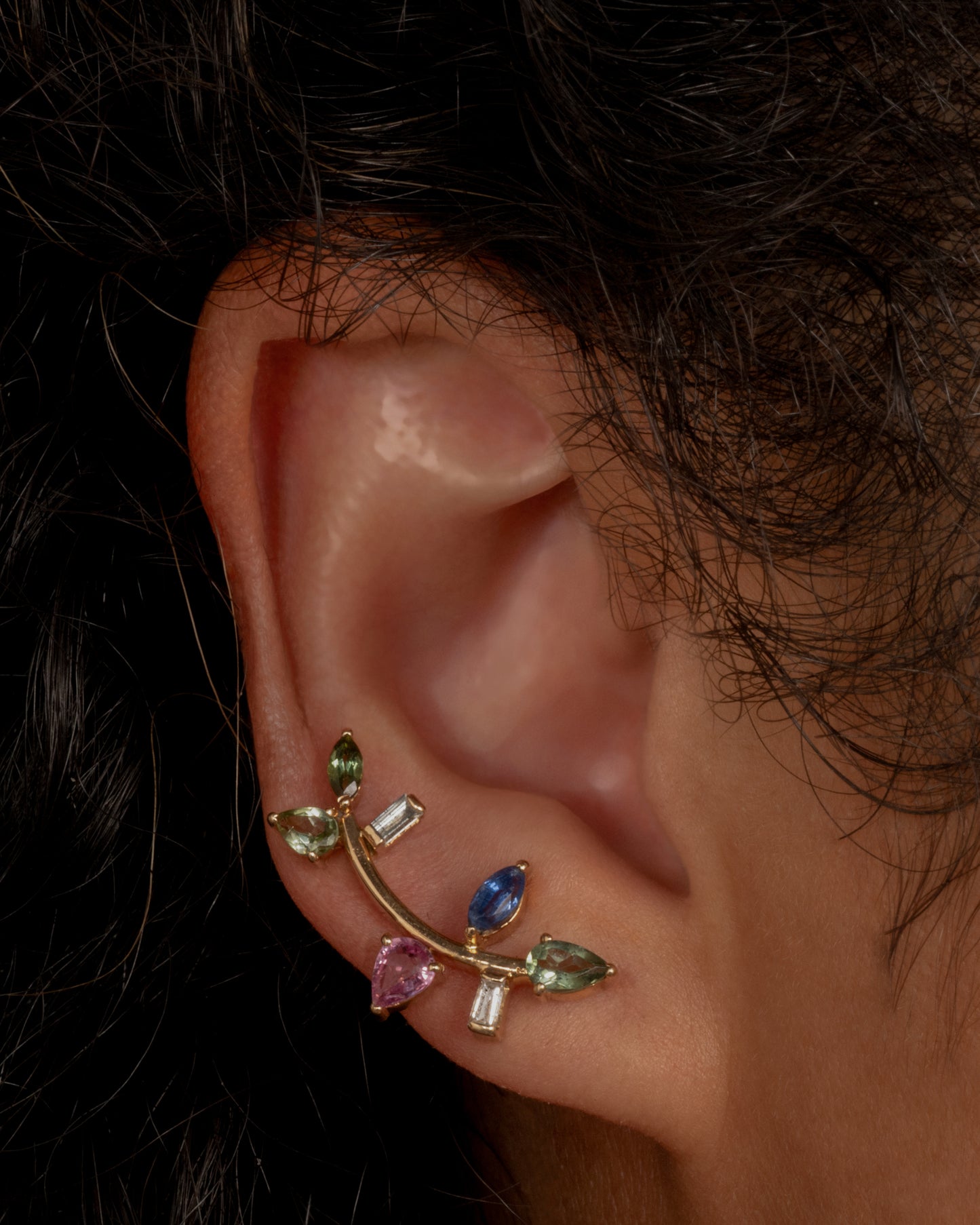 This elegant 14k gold stud, dotted with blue and green gemstones, reminds us of an olive branch. Shown worn in an ear.