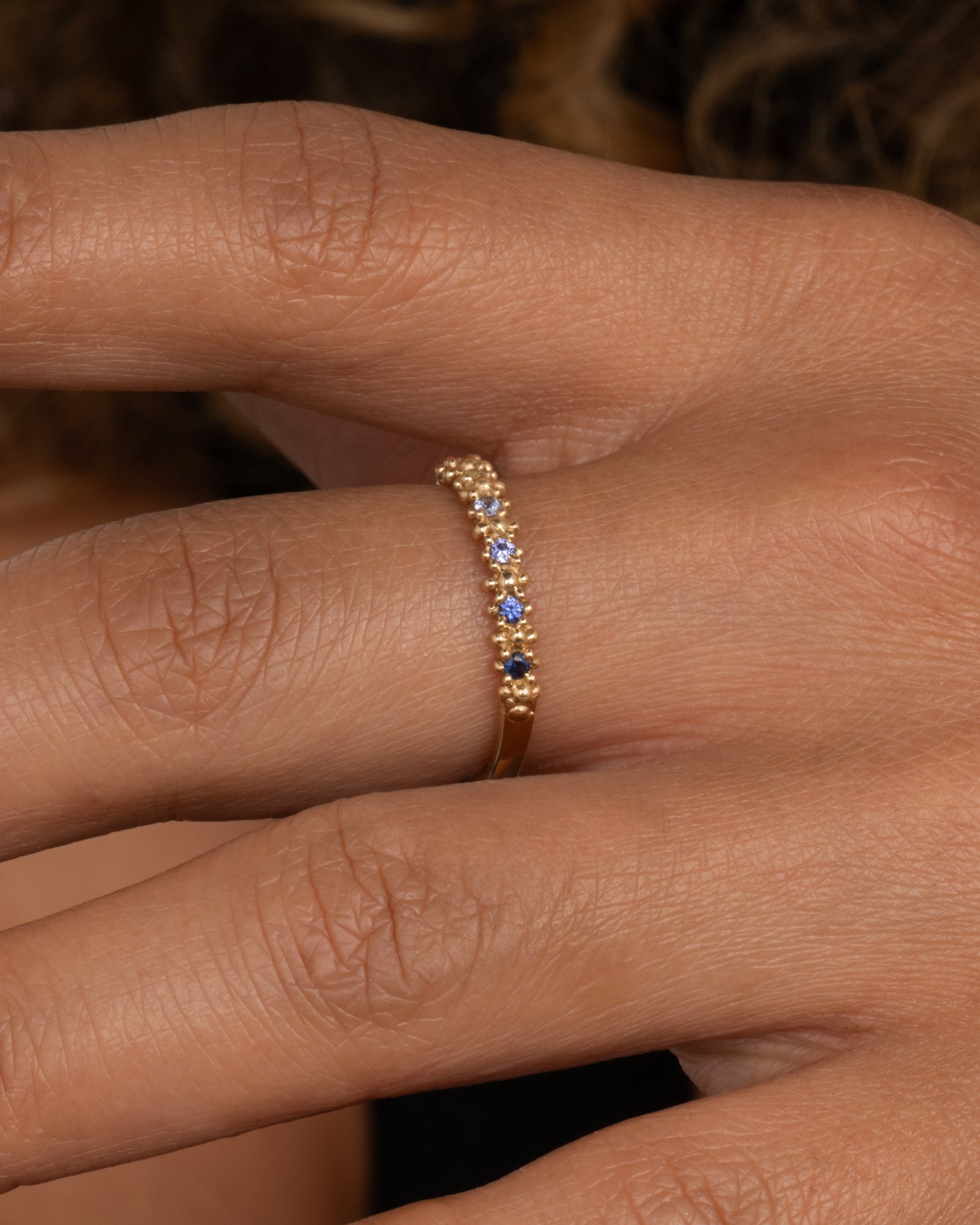 A textured yellow gold band with an ombré queue of six sapphires shown from the side on a hand.