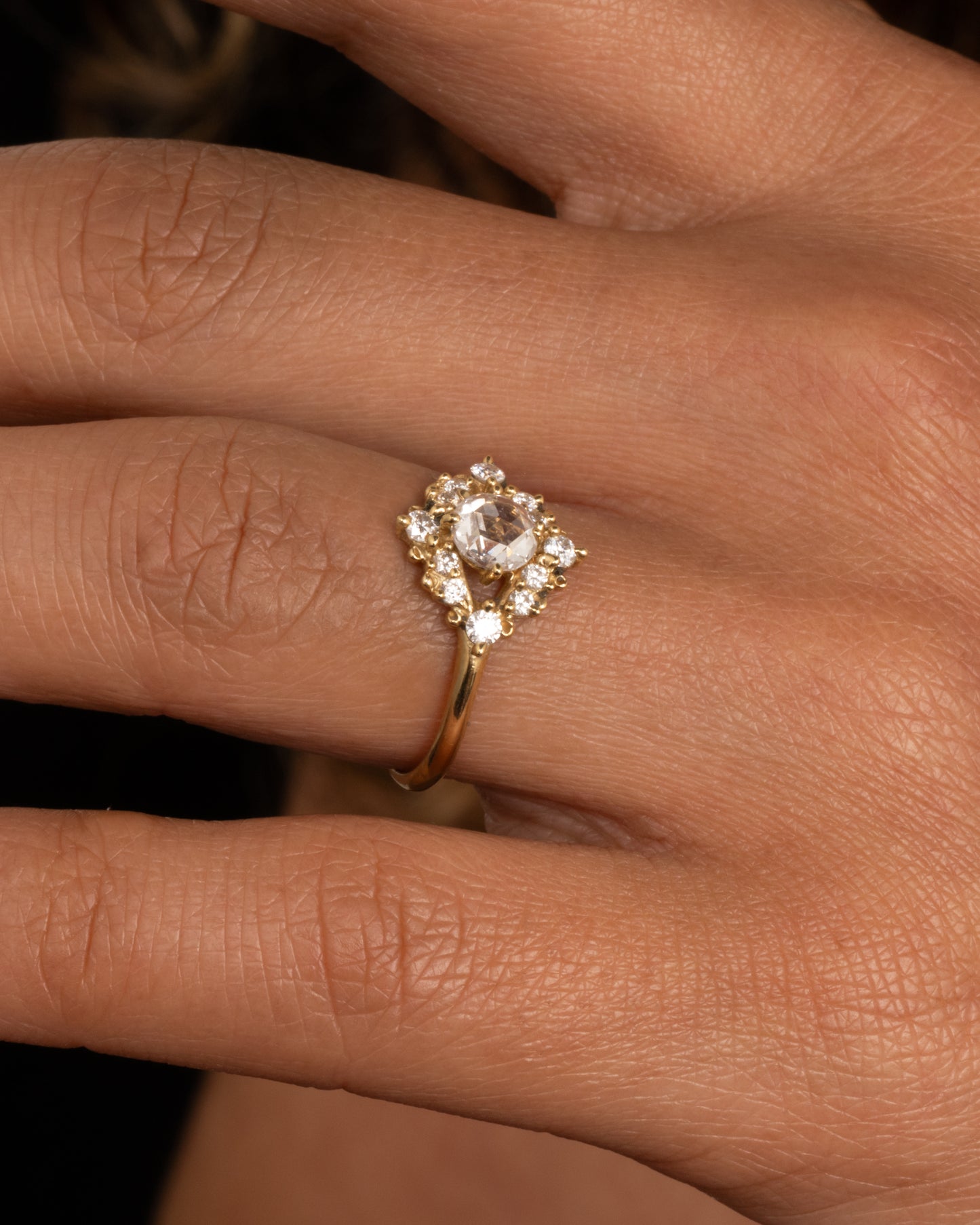 A yellow gold ring with a rose cut center diamond surrounded by round diamonds. Shown worn on hand.