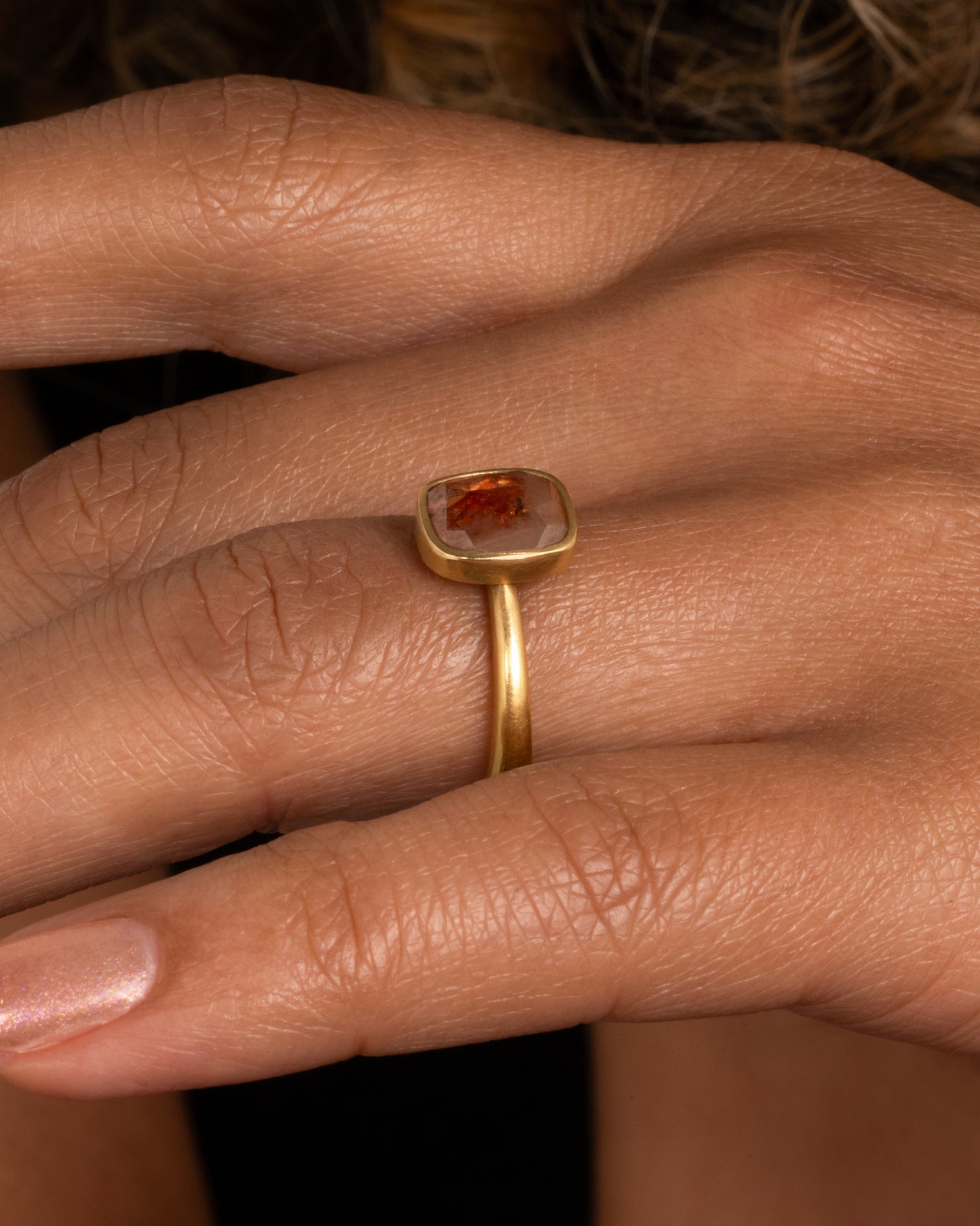A yellow gold ring with a cushion shaped, rusty-red diamond. Shown from the side on a hand.