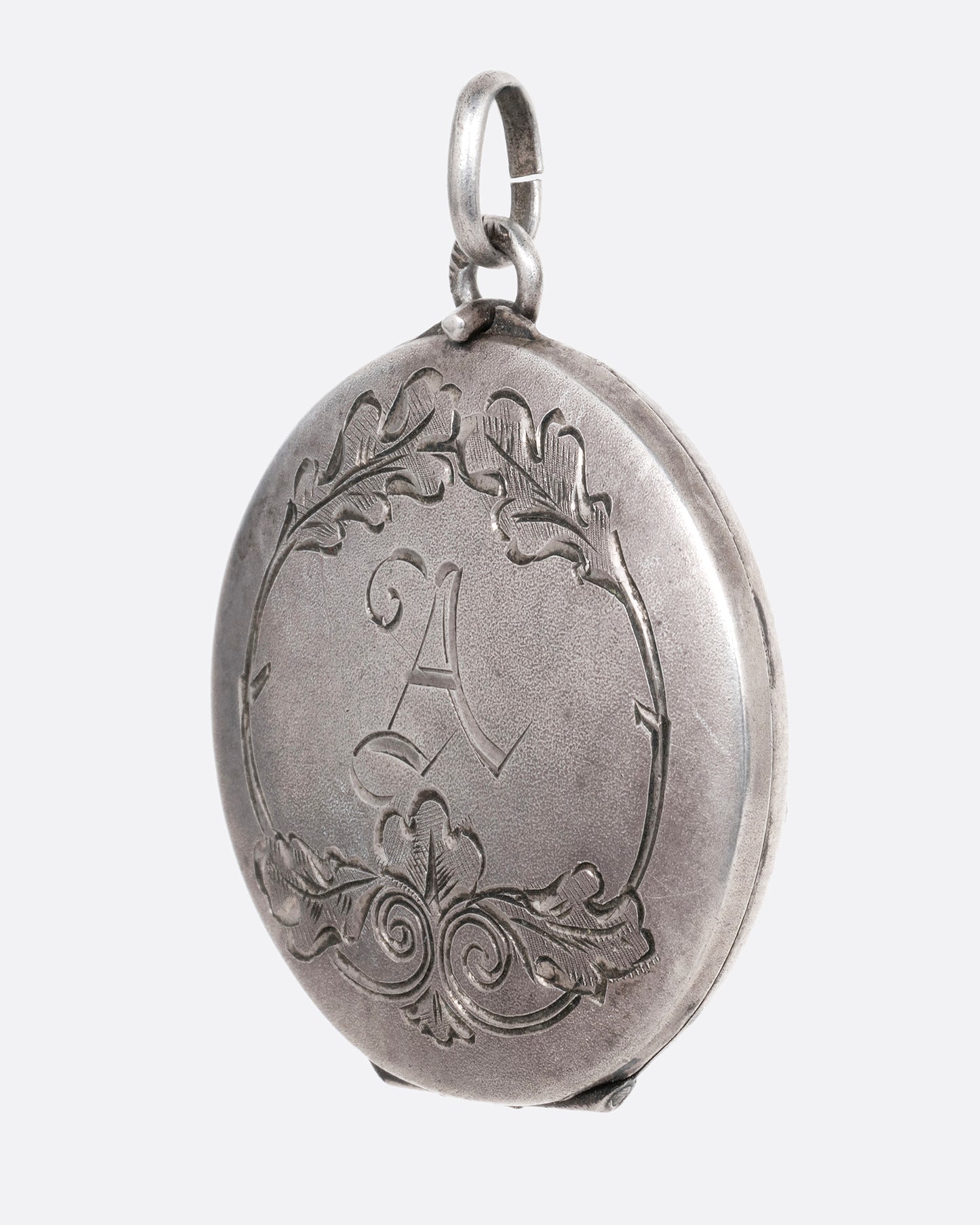 A silver locket with an engraved floral motif and an A' initial at the center. Shown from the side.