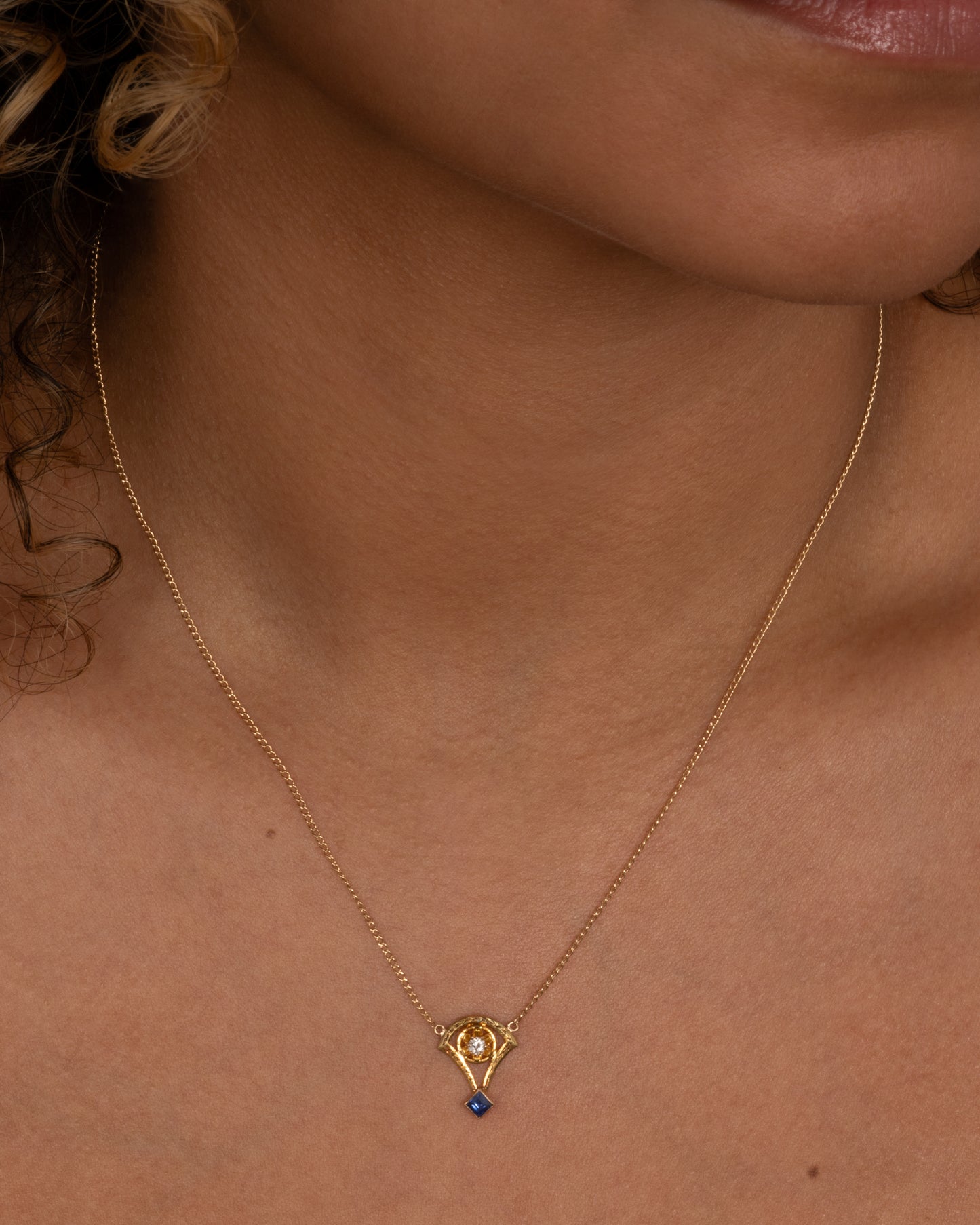 A yellow gold cable chain necklace with a pendant shaped like the basket of a mini hot air balloon with a prong set diamond. and princess cut sapphire. Shown from the front on a person.