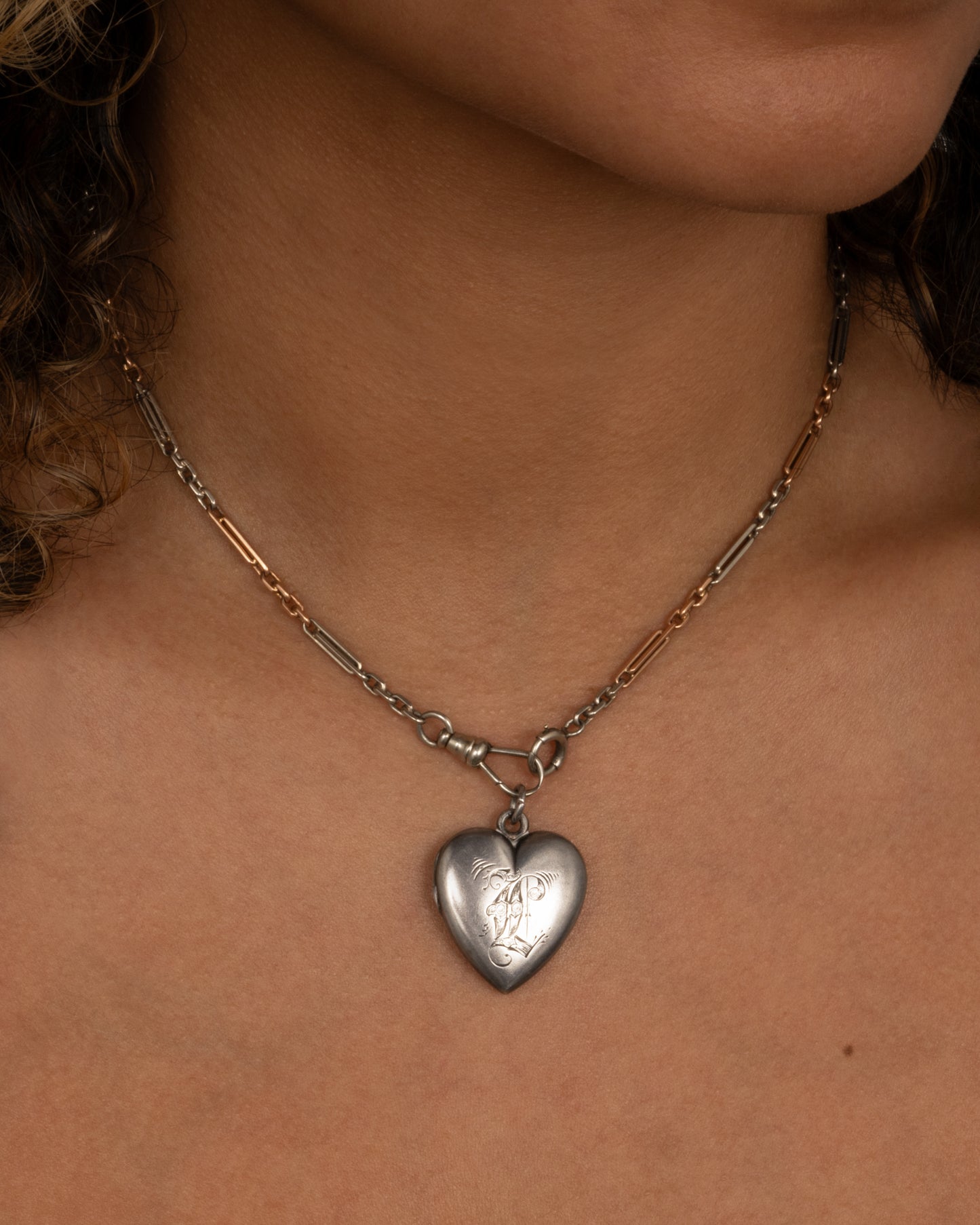 A silver heart shaped locket with an engraved 'L'. Shown on a chain on a person from the front.