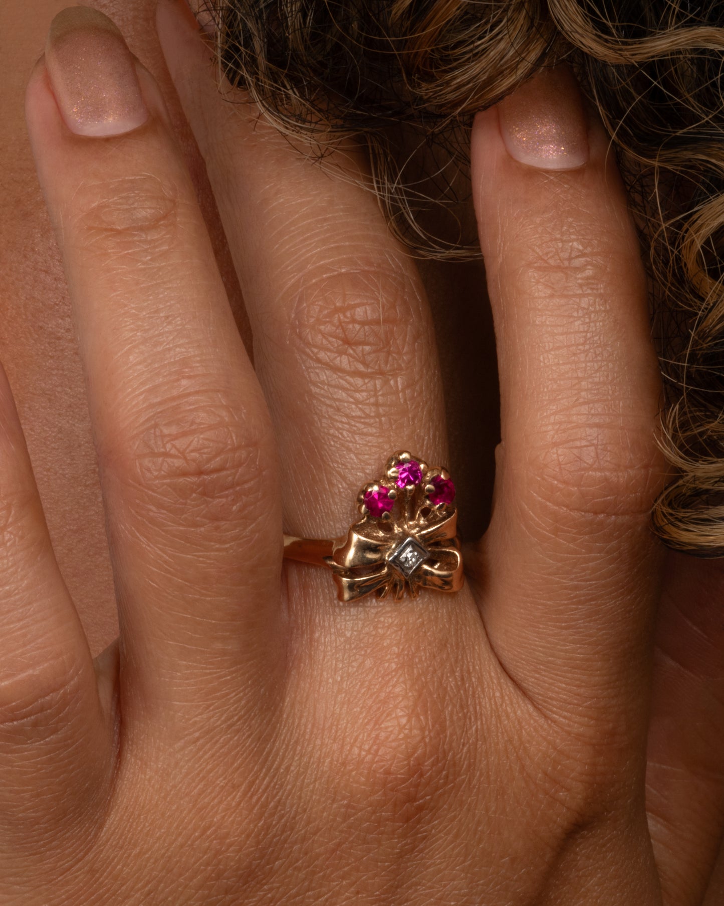 A yellow gold ring with a bouquet of ruby topped stems tied in a ribbon-like gold band and topped with a princess cut diamond. Shown from the front on a person.