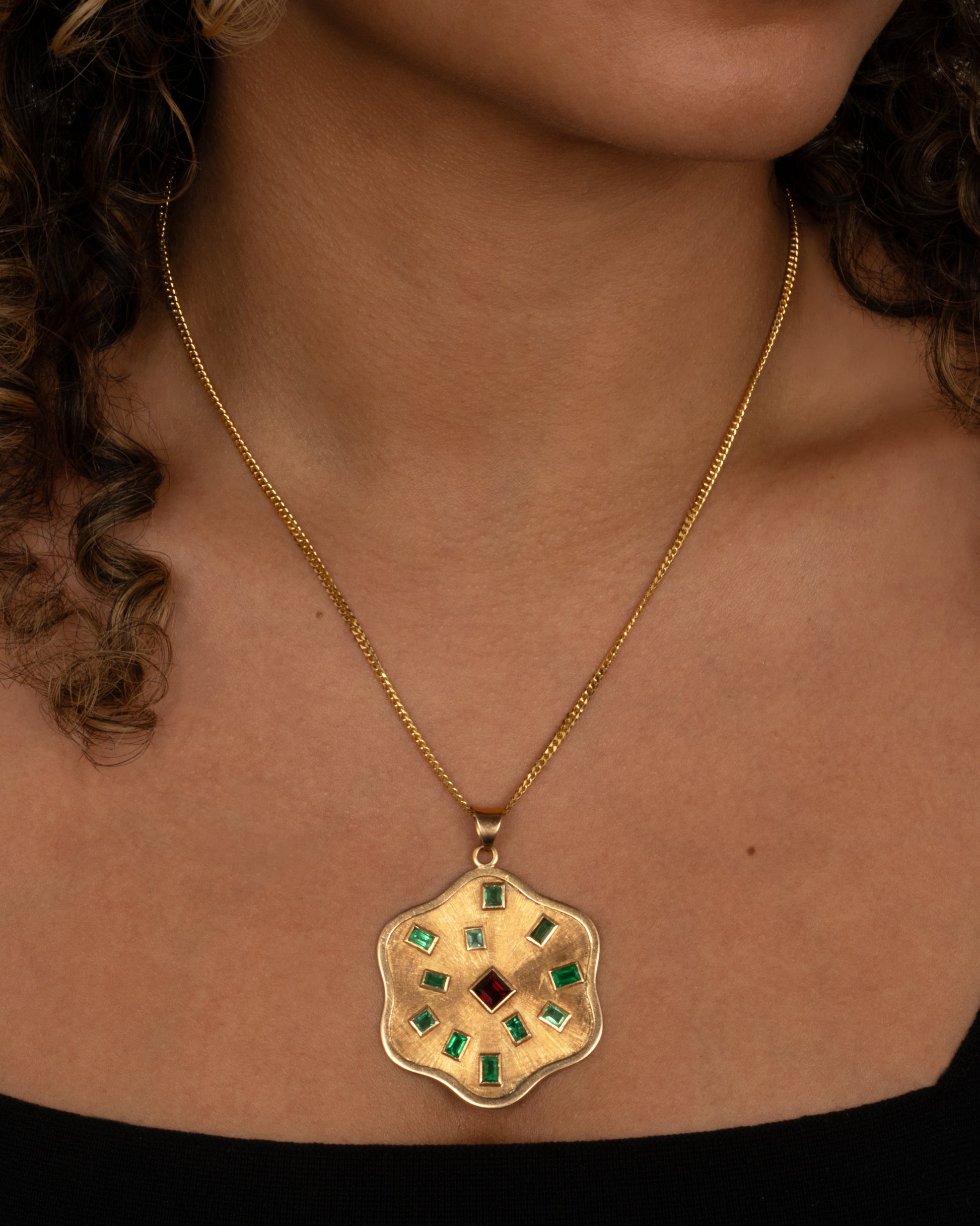 A yellow gold wavy edged medallion pendant with baguette emeralds encircling a central princess cut garnet. Shown from the front on a person.
