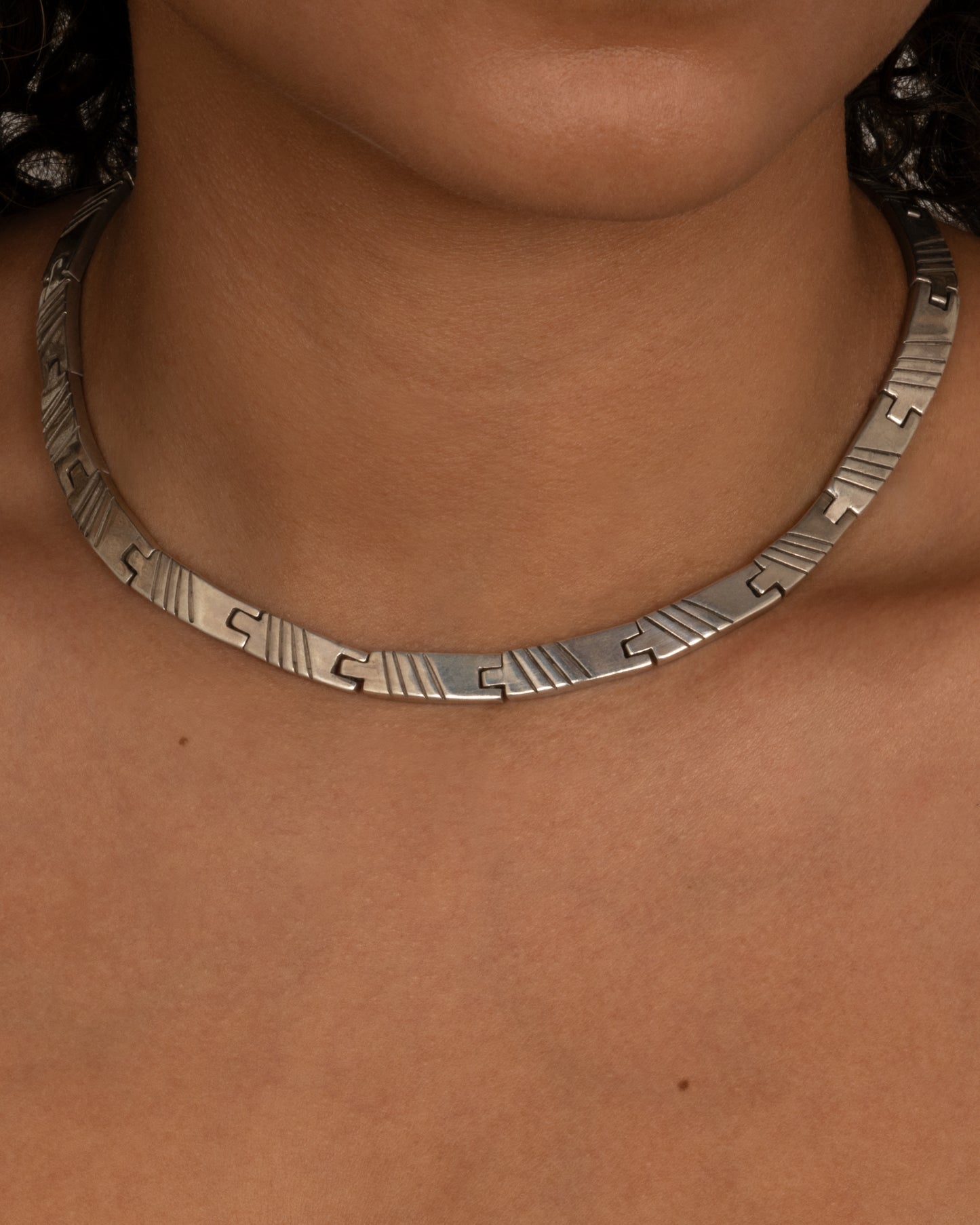 A silver collar necklace with hinged rectangular links and striped etchings. Shown from the front on a person.