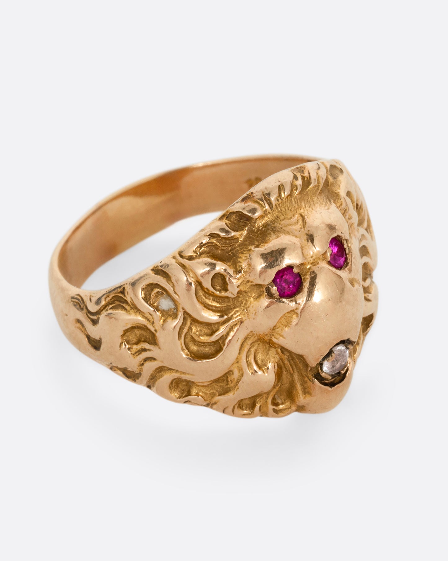 A yellow gold lion ring with ruby eyes and a single diamond in its mouth. Shown from the side.