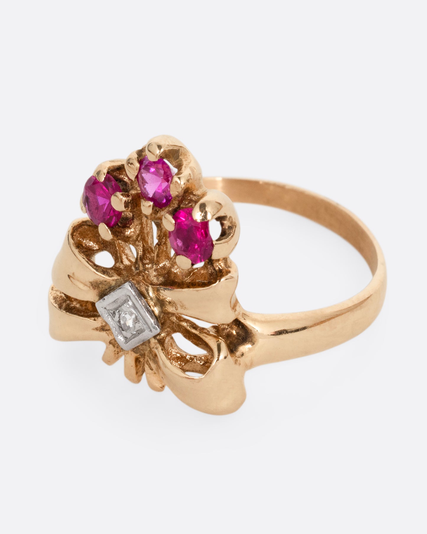 A yellow gold ring with a bouquet of ruby topped stems tied in a ribbon-like gold band and topped with a princess cut diamond. Shown from the side.