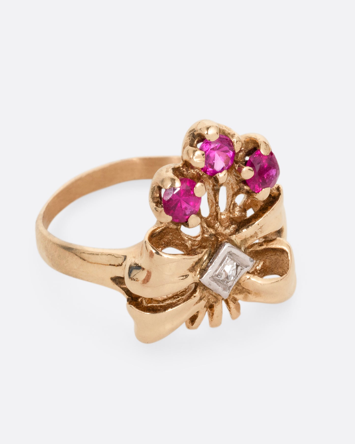 A yellow gold ring with a bouquet of ruby topped stems tied in a ribbon-like gold band and topped with a princess cut diamond. Shown from the side.