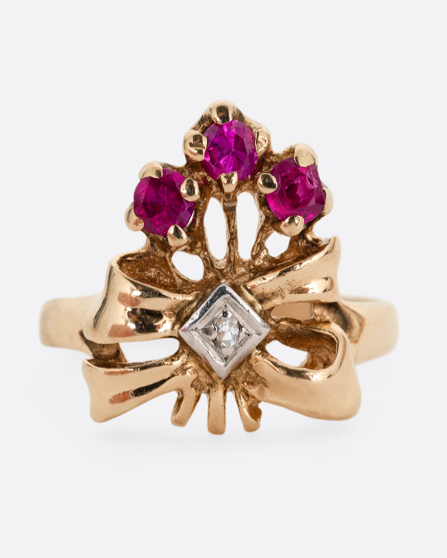 A yellow gold ring with a bouquet of ruby topped stems tied in a ribbon-like gold band and topped with a princess cut diamond. Shown from the front.