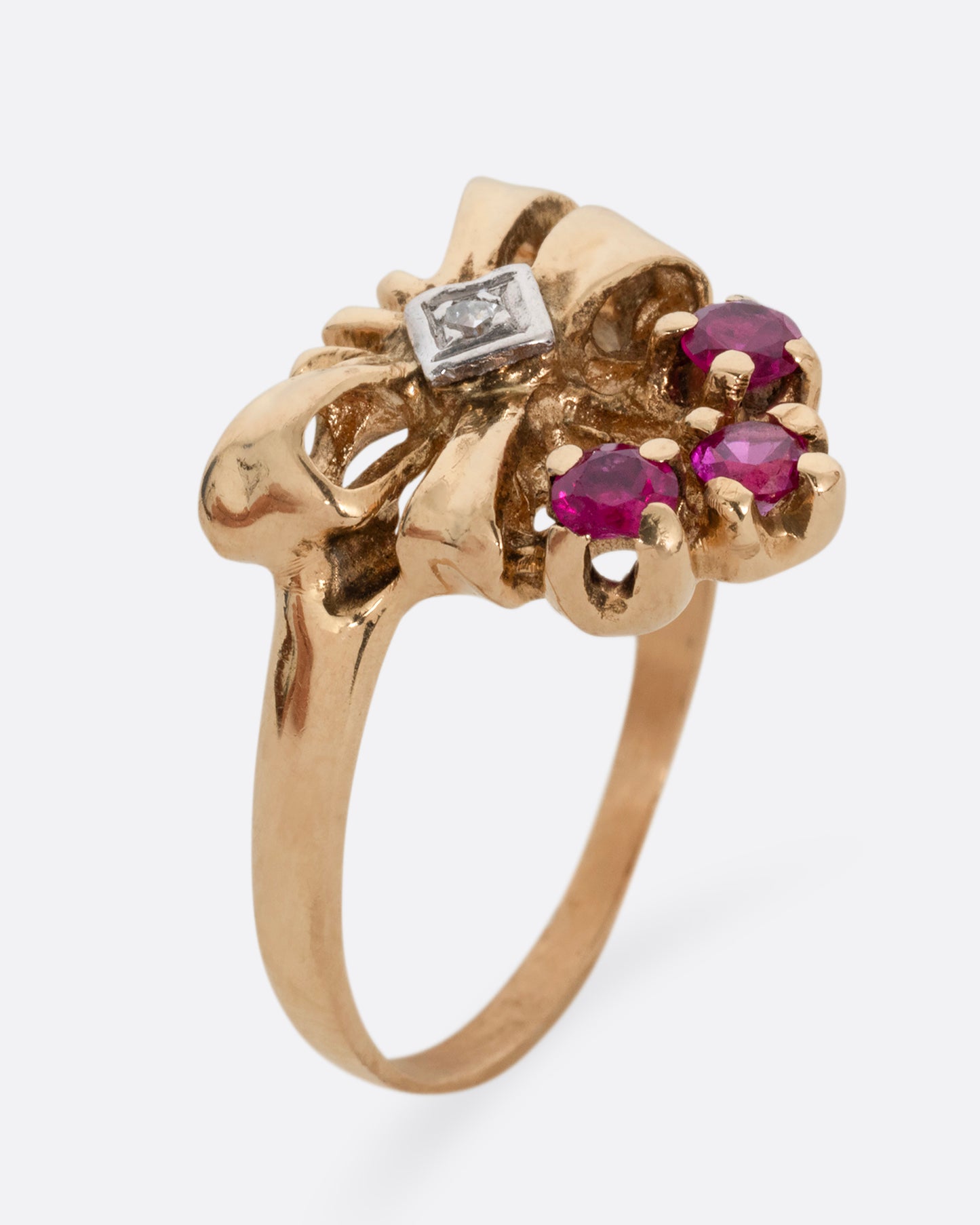 A yellow gold ring with a bouquet of ruby topped stems tied in a ribbon-like gold band and topped with a princess cut diamond. Shown standing up from the side.