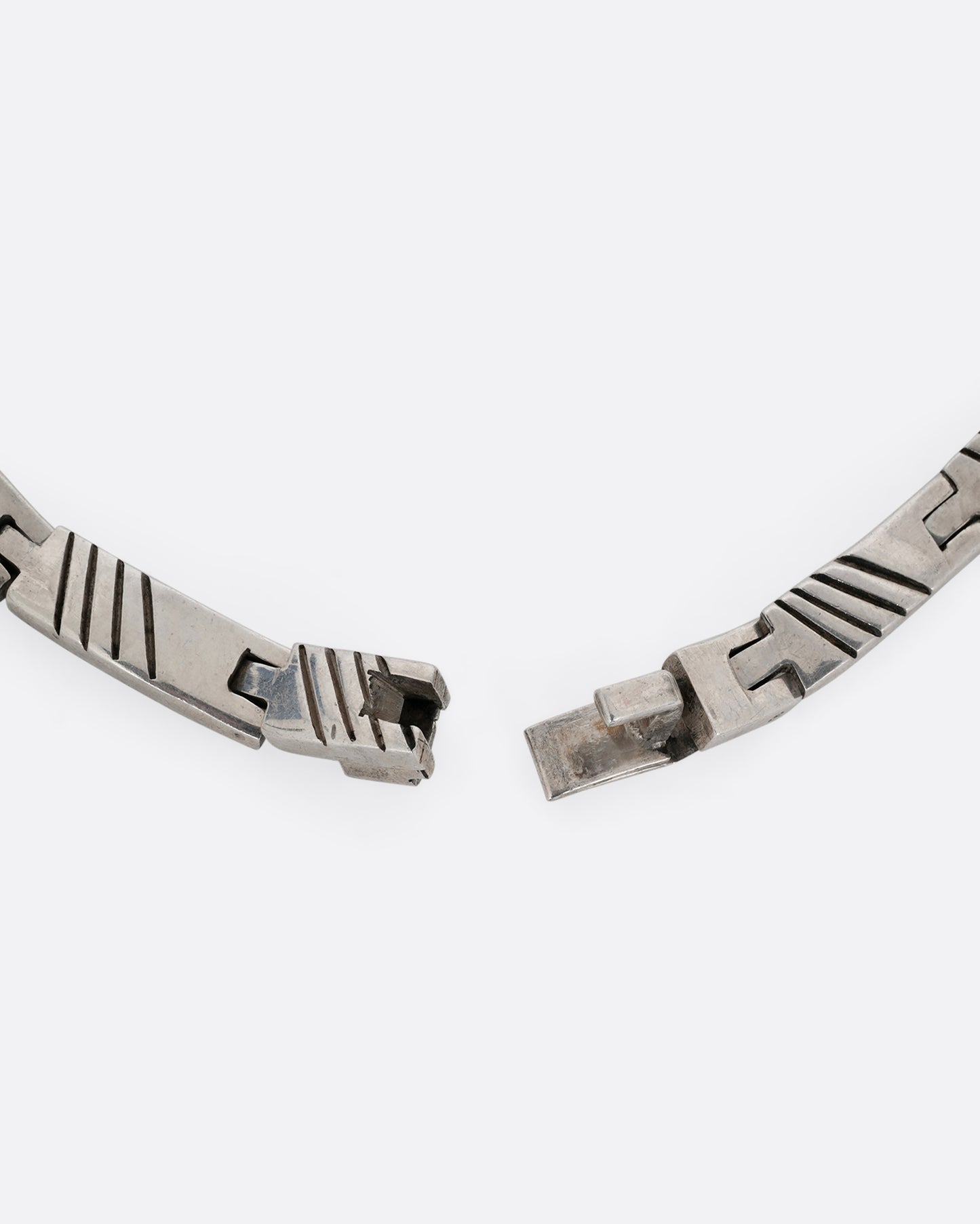 A close up of the clasp of a silver collar necklace with hinged rectangular links and striped etchings.