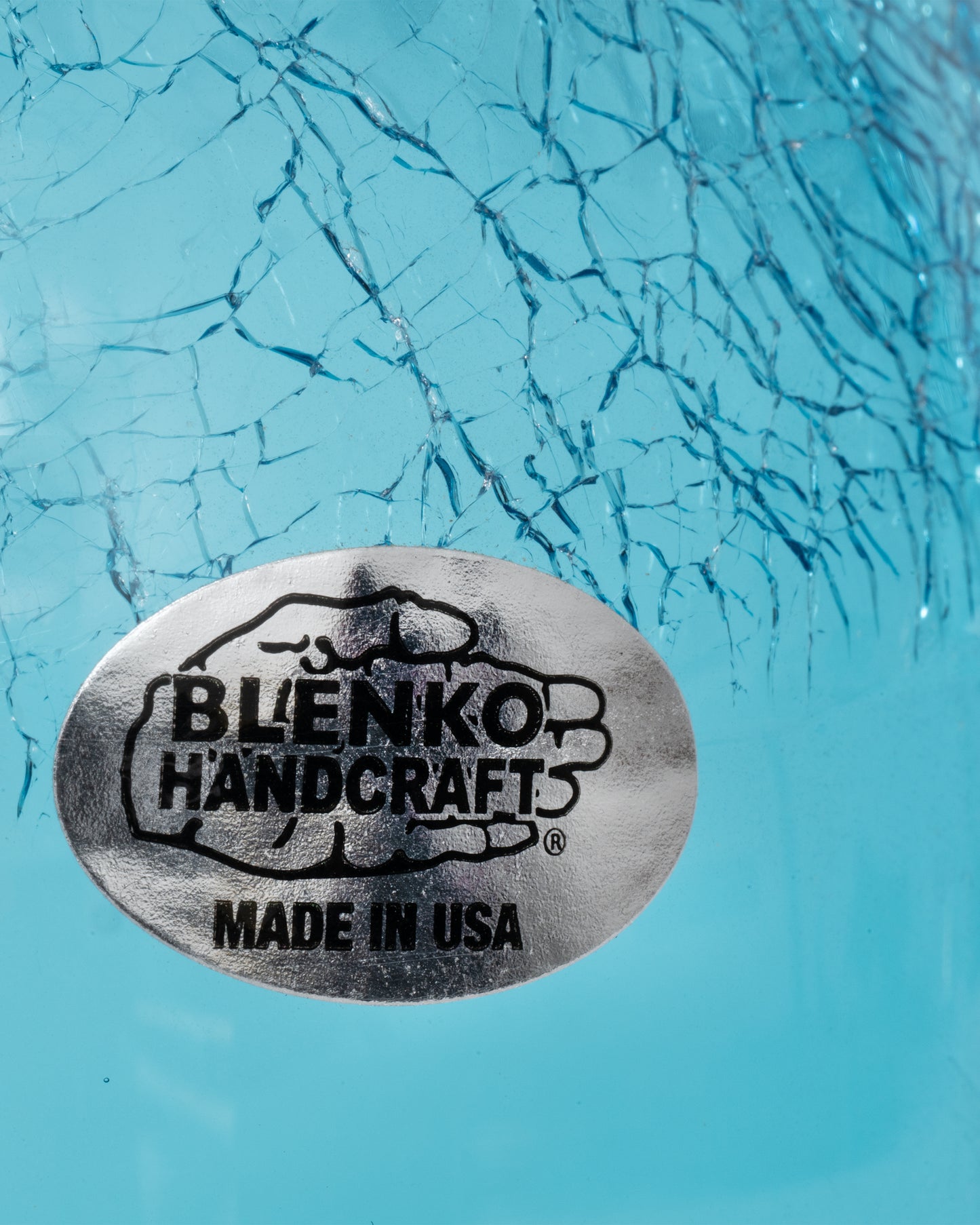 A close up of the Blenko sticker on a A glass aqua moonshine jug with a clear crystal handle, a crackled finish at the top, and a cork stopper.