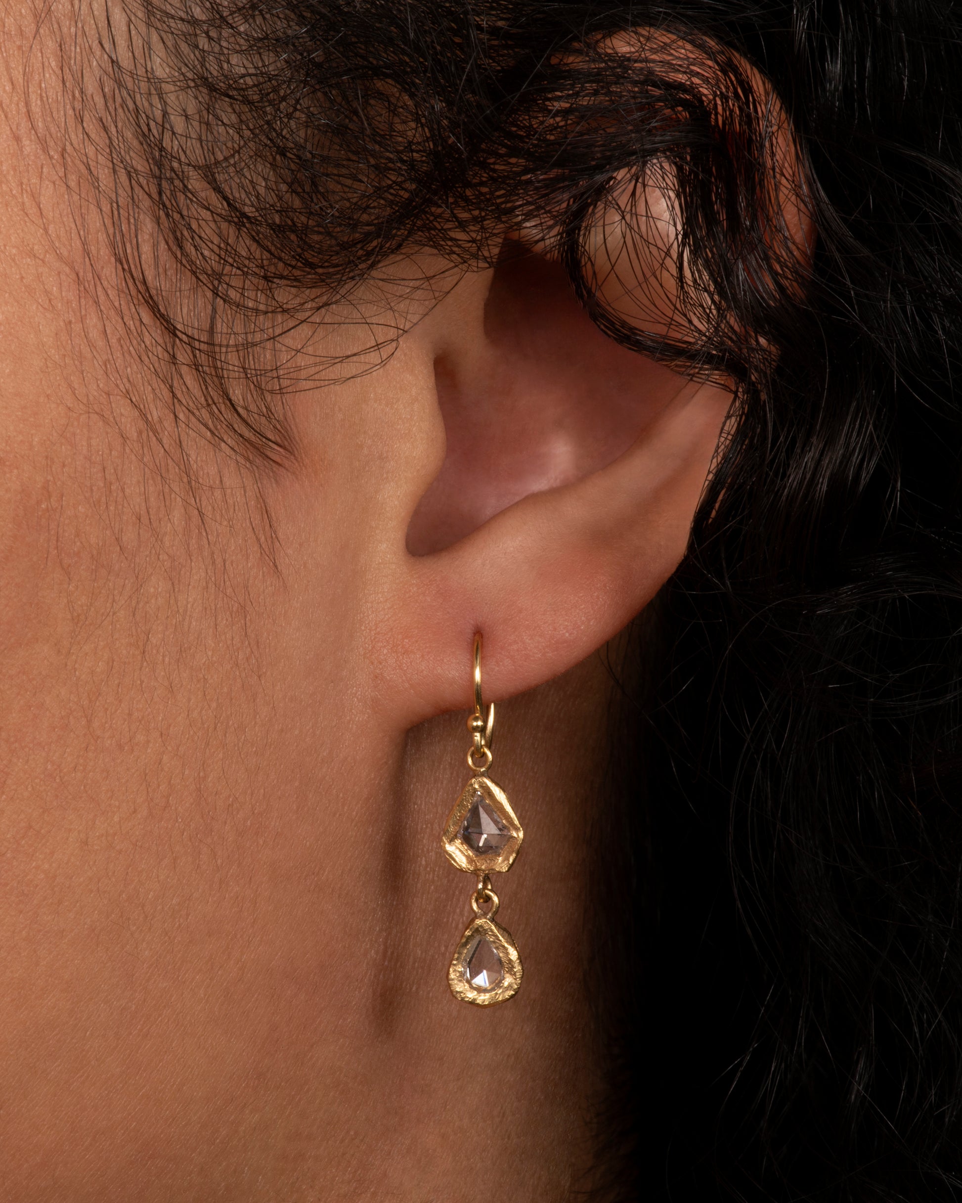A slightly asymmetrical yellow gold diamond drop earring with two diamonds set in hand carved bezels. Shown from the front on a person.