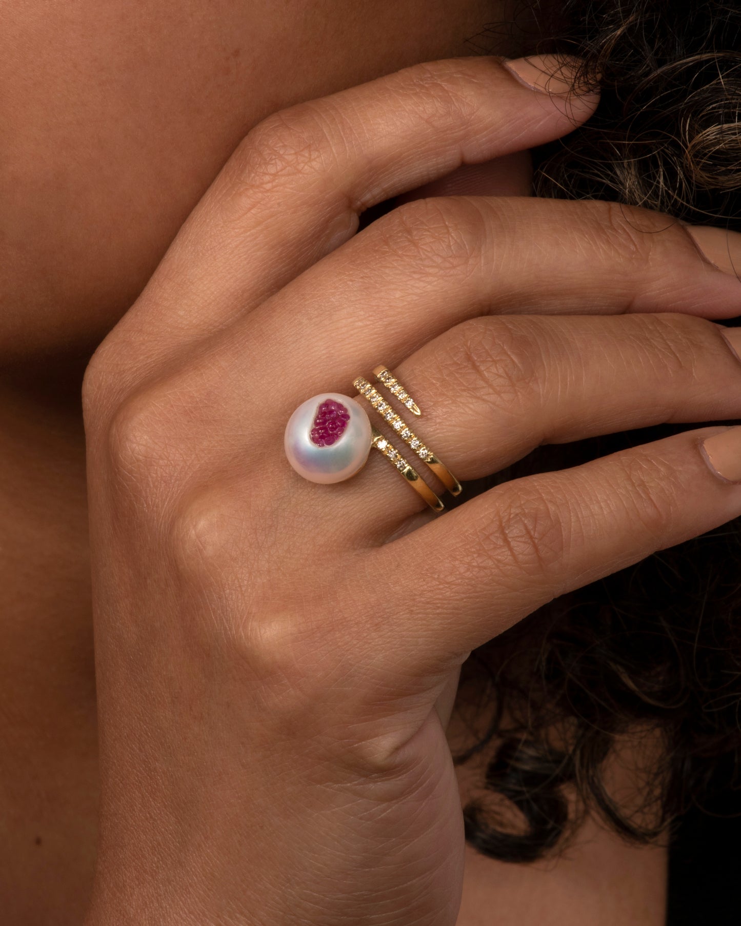 A yellow gold wrap ring with an organic freshwater pearl lined with rubies. Shown from the front on a hand.