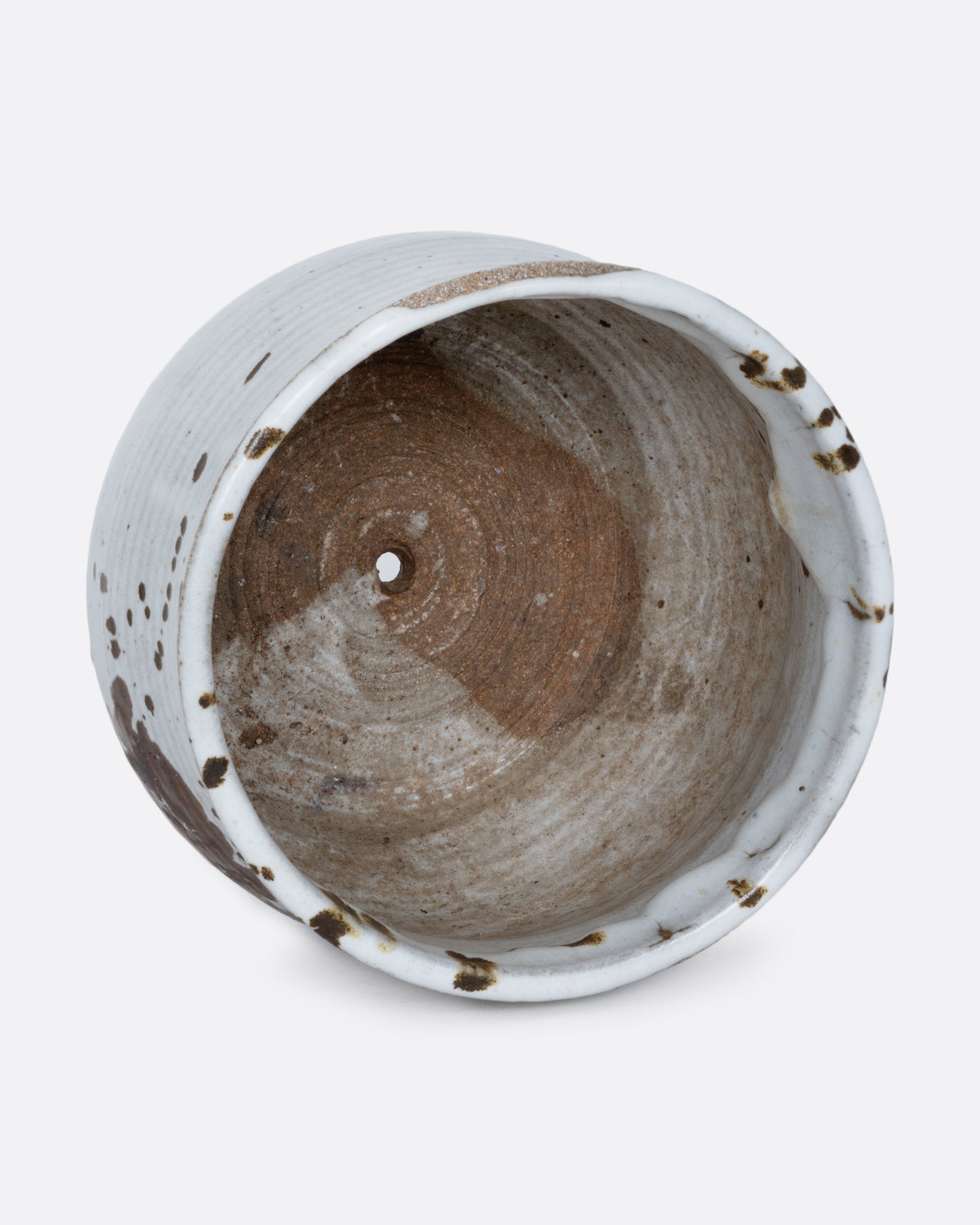 A white pot with a brown splattered glaze and a drainage hole at its base. Shown lying on its side from the top.