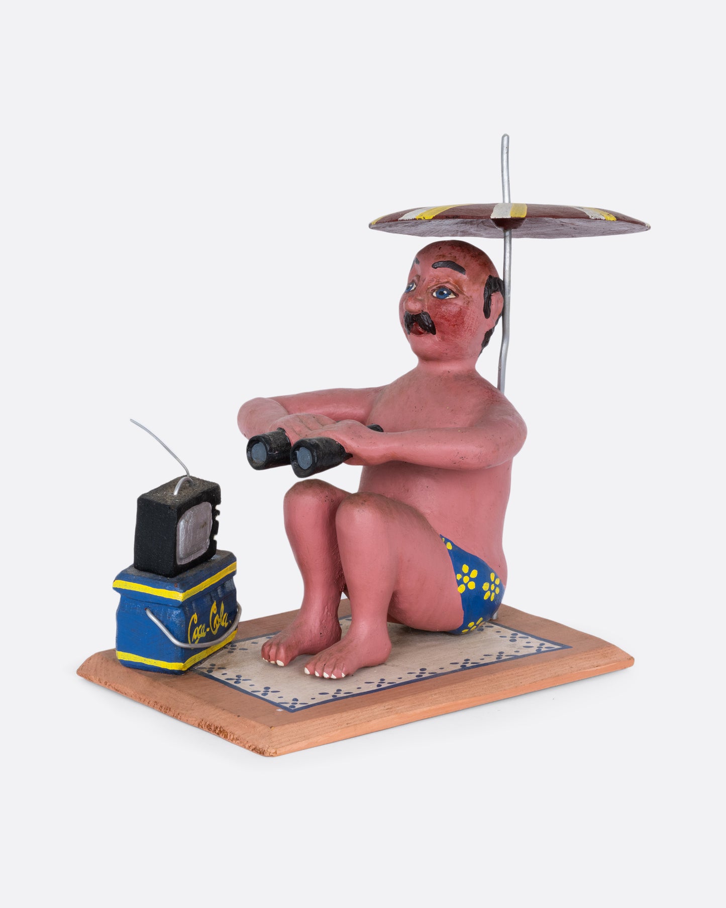 A hand carved and painted wooden sculpture of a beach goer sitting with an umbrella, tv, cooler, and binoculars. Shown from the side.
