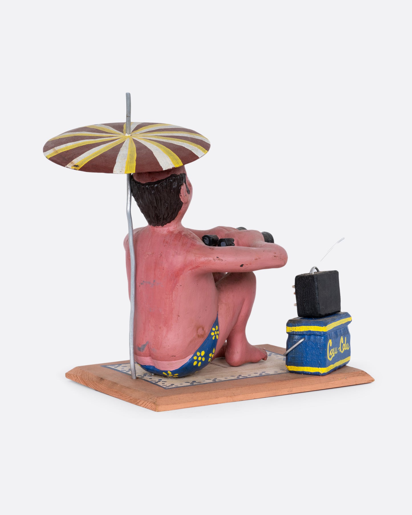 A hand carved and painted wooden sculpture of a beach goer sitting with an umbrella, tv, cooler, and binoculars. Shown from the back.