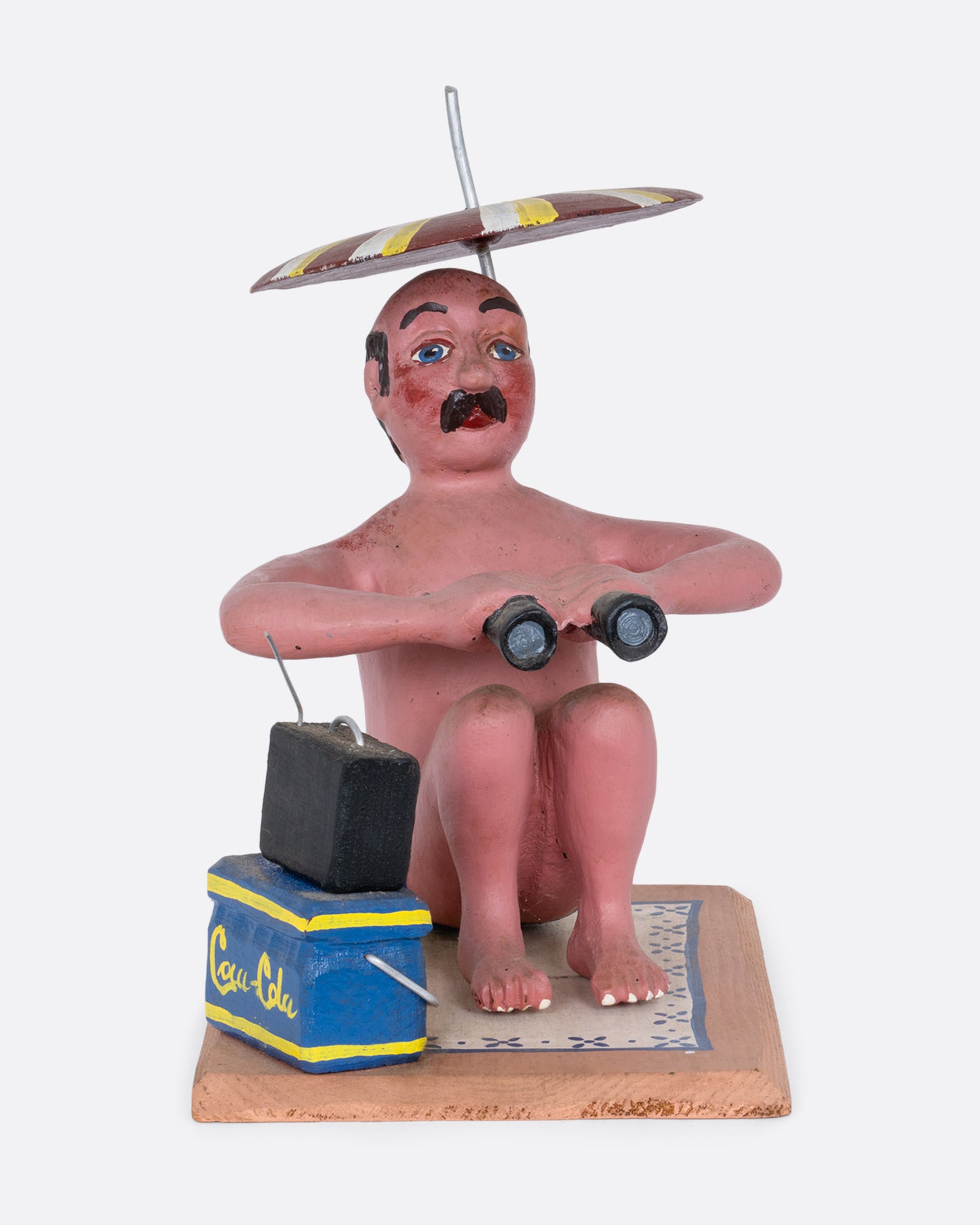 A hand carved and painted wooden sculpture of a beach goer sitting with an umbrella, tv, cooler, and binoculars. Shown from the front.