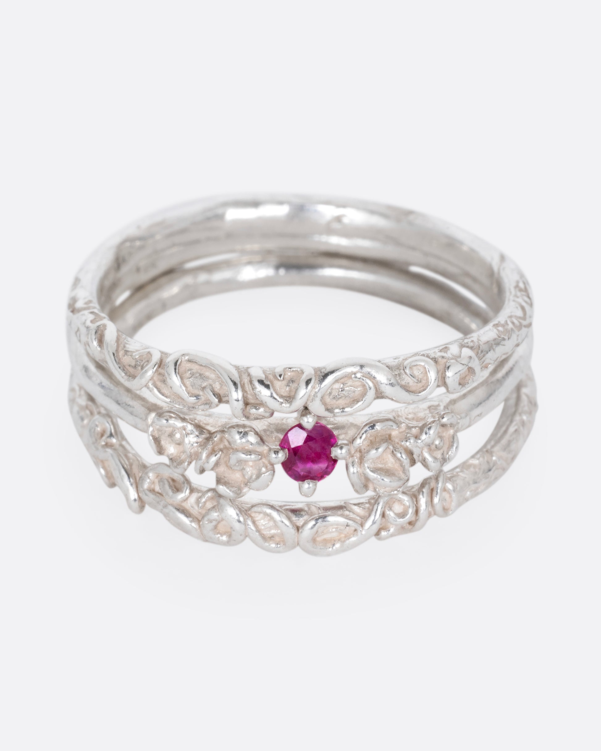A silver ring with three bands and a round prong set ruby at the center, shown from the front.